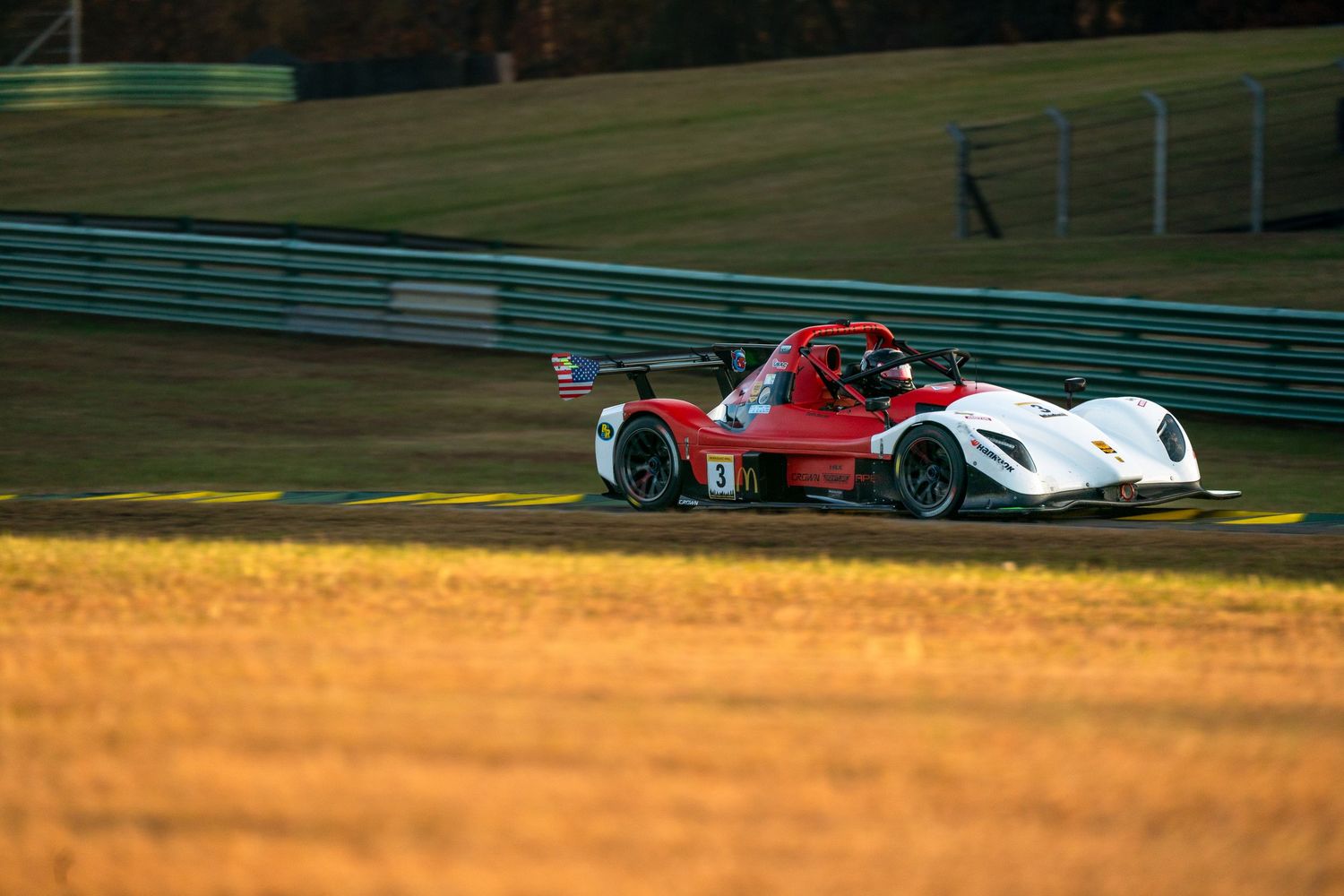 Red and white race cars speed along a racetrack at sunset with golden light illuminating the grass.