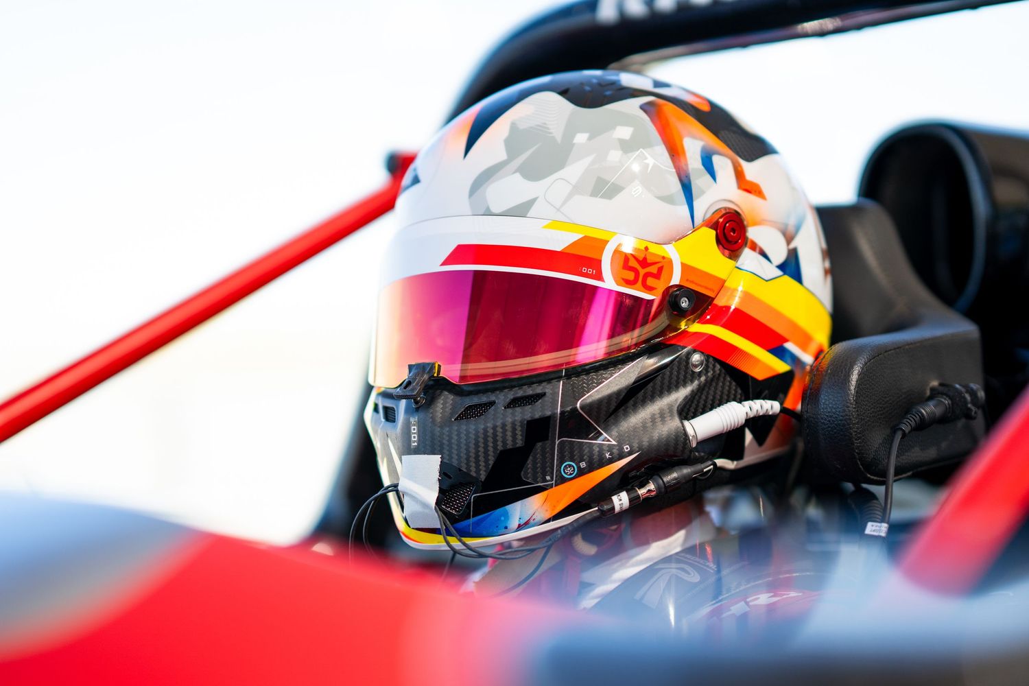 Side view of race car driver in cockpit with distinctive orange and blue helmet design.