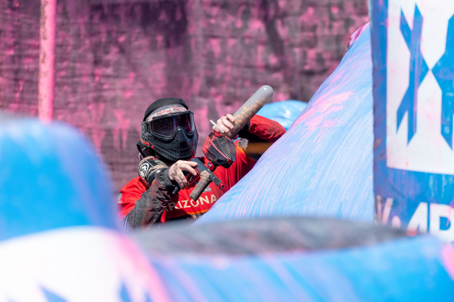 Paintball player in red Arizona jersey leaning over a blue inflatable bunker during competition.
