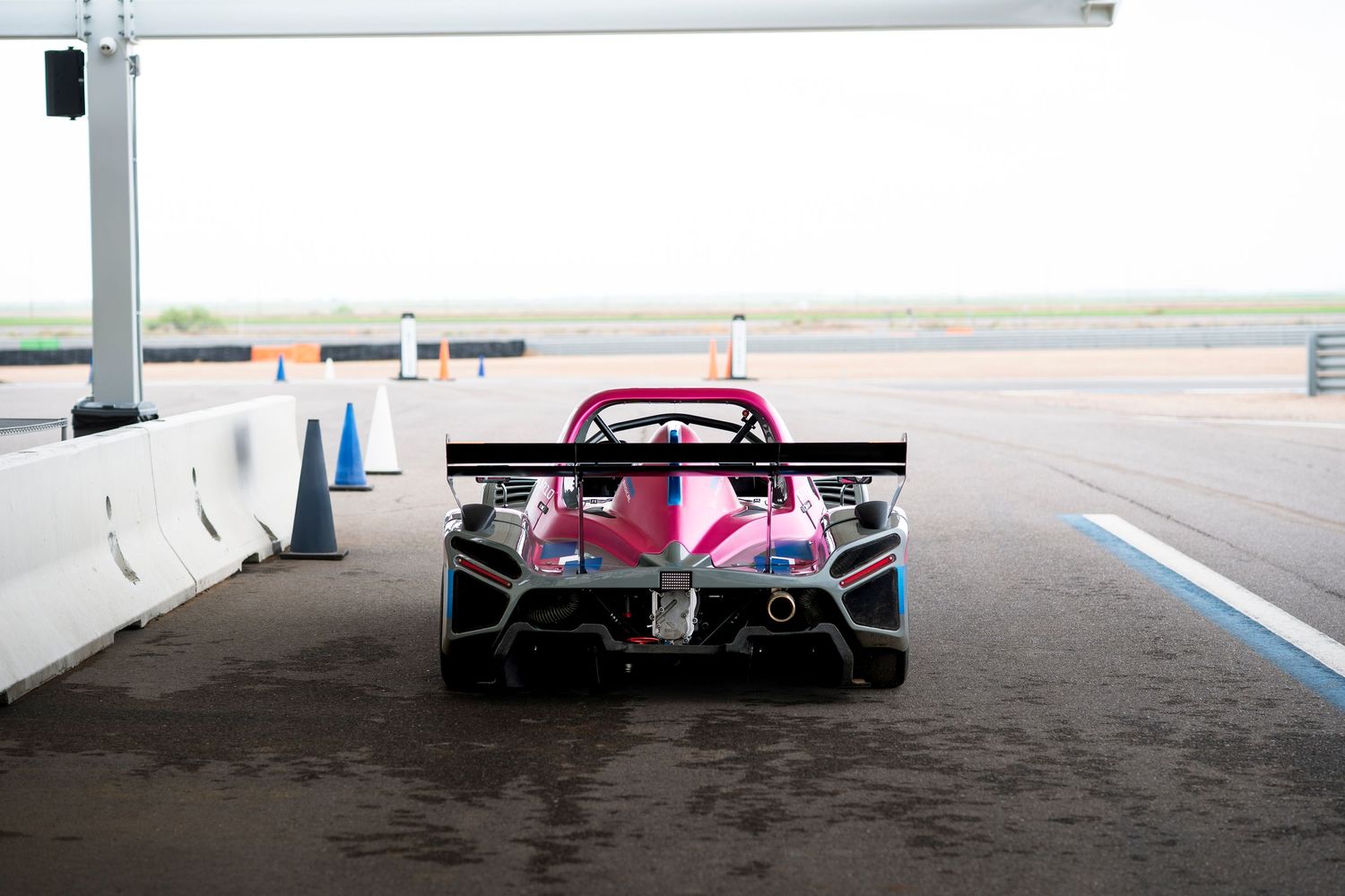 Rear view of a sleek pink and silver race car with large rear wing parked in a pit lane.
