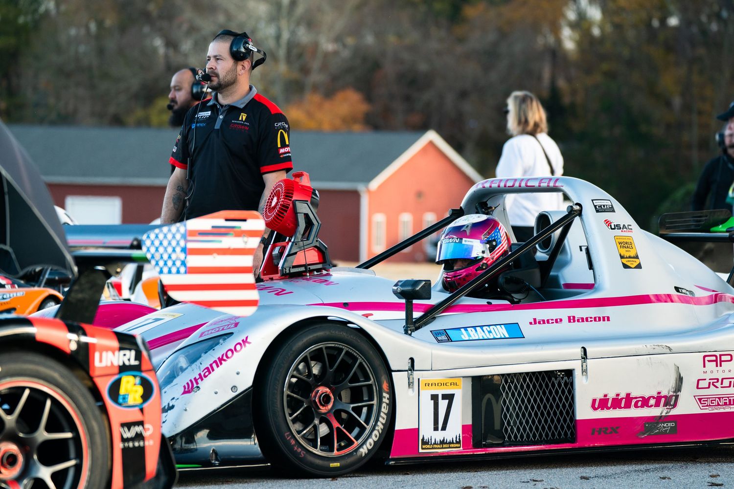 Row of Formula race cars lined up on track with American flags visible in background.