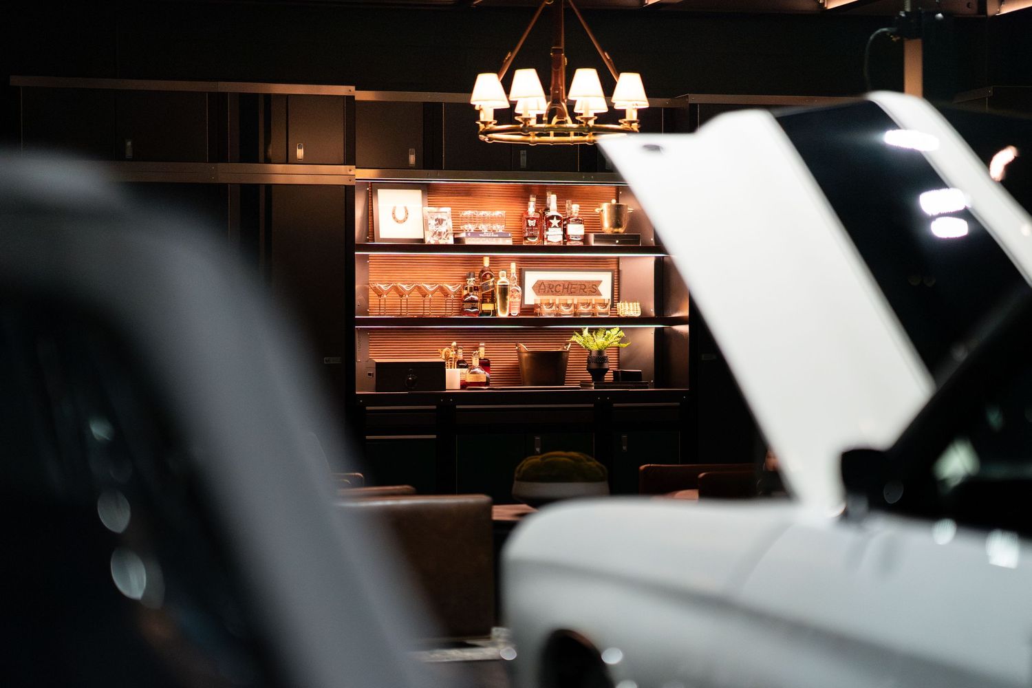 Dimly lit bar interior with open car hood in foreground and illuminated shelving with bottles in background.