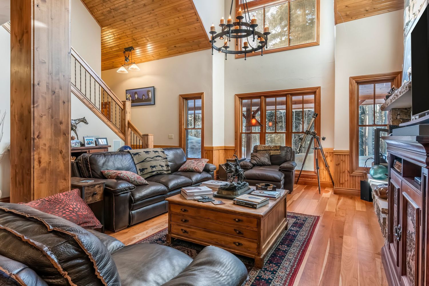 Spacious lodge room with wooden staircase, leather furniture, and cathedral ceiling with exposed beams.