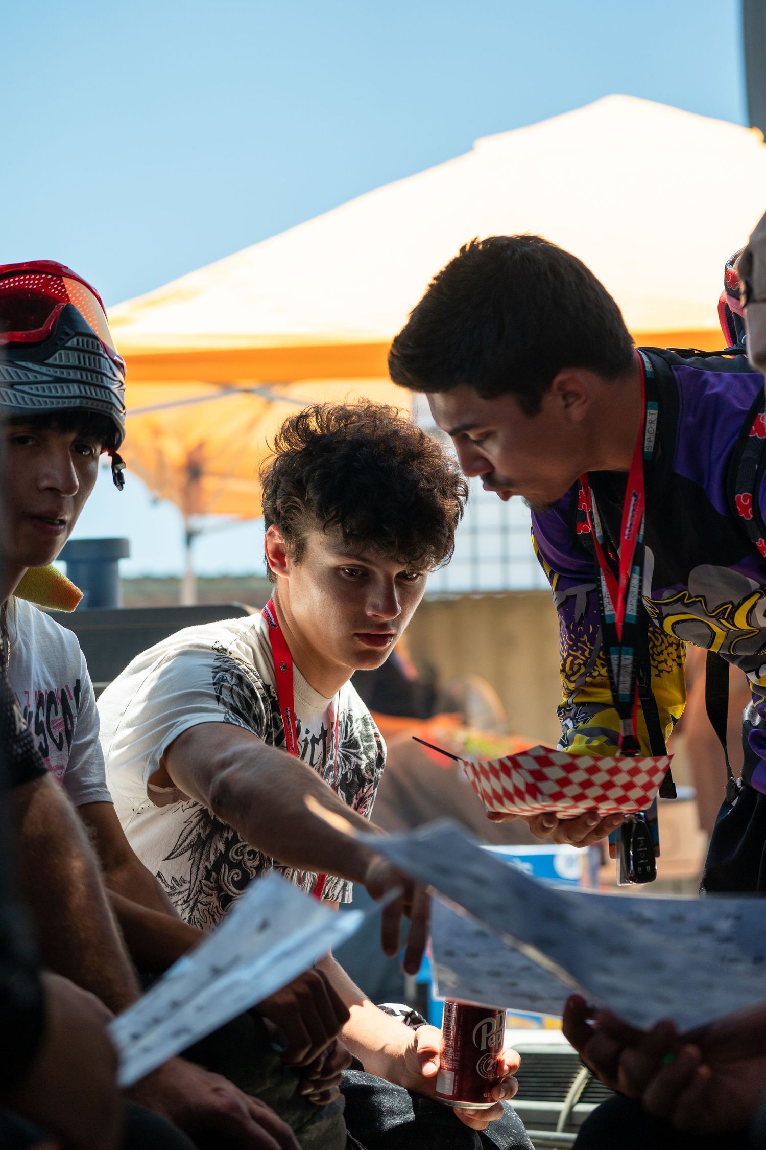 Three young men reviewing documents at an outdoor event under a yellow umbrella on a sunny day.
