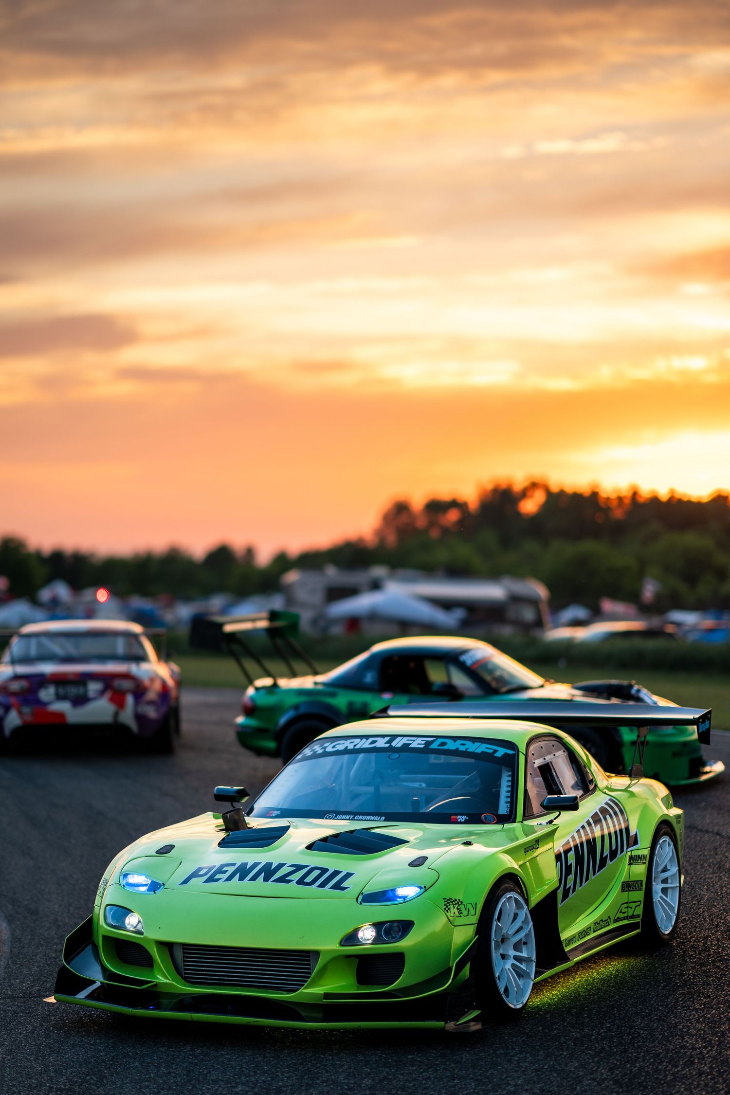 Green Pennzoil Mazda RX-7 race car on track at sunset with other racing cars in background.