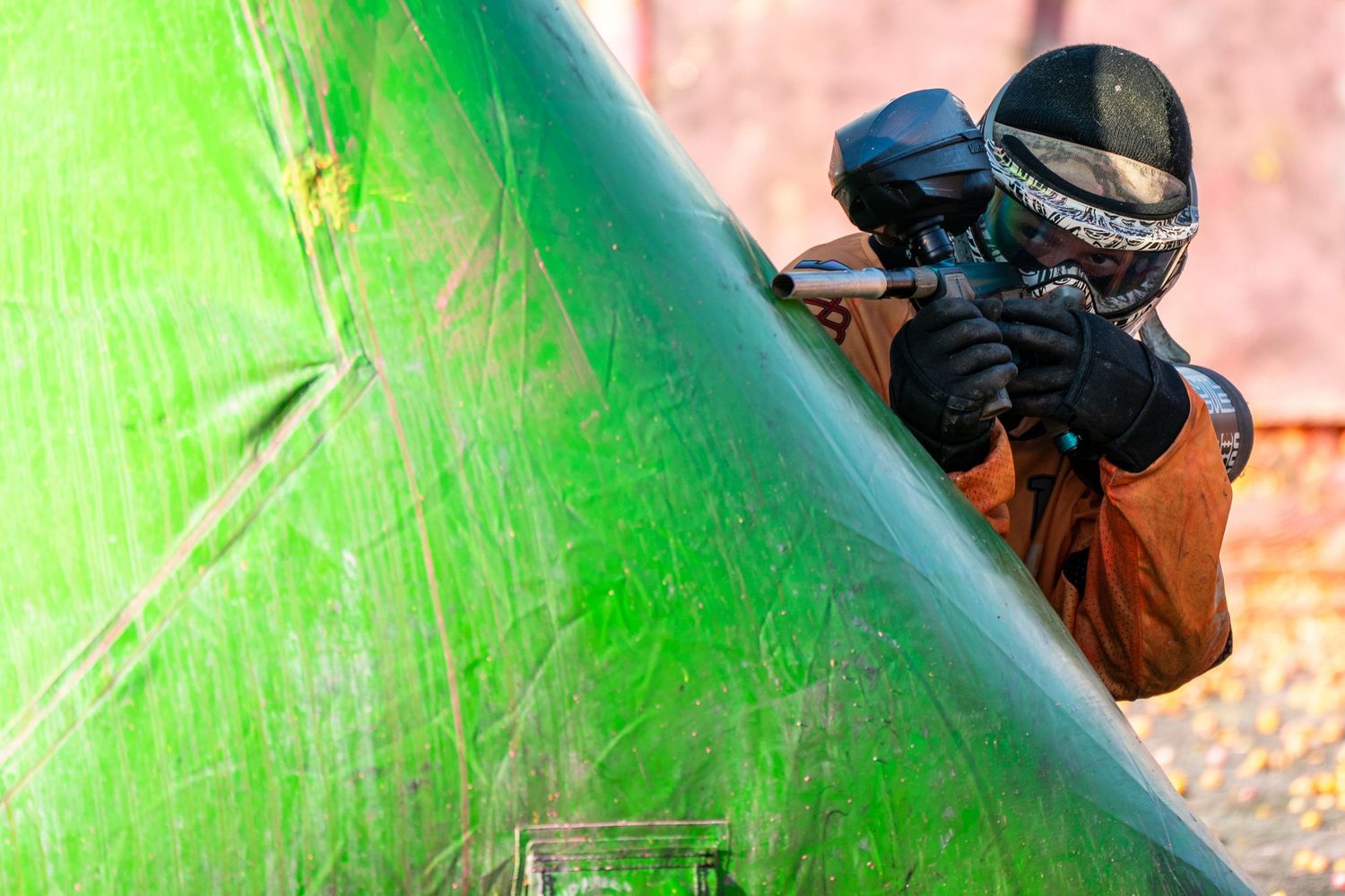Paintball player in protective mask aiming marker while taking cover behind bright green inflatable bunker.