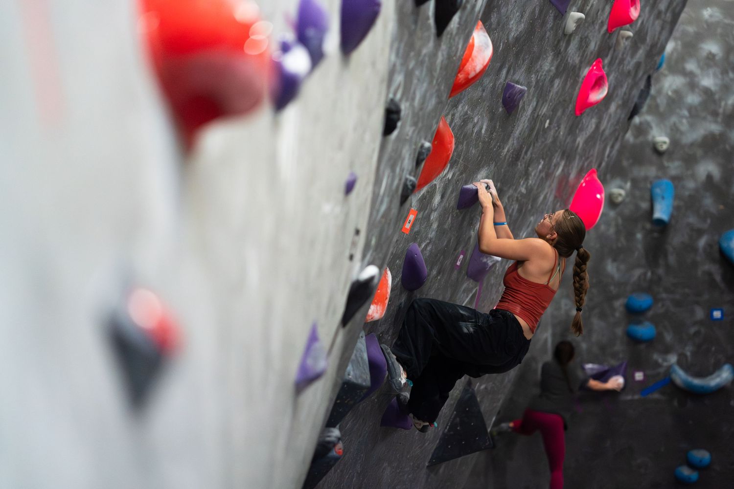 Climber in a red tank top and black pants reaches for a hold on an indoor bouldering wall covered in colorful red, purple and pink grips.