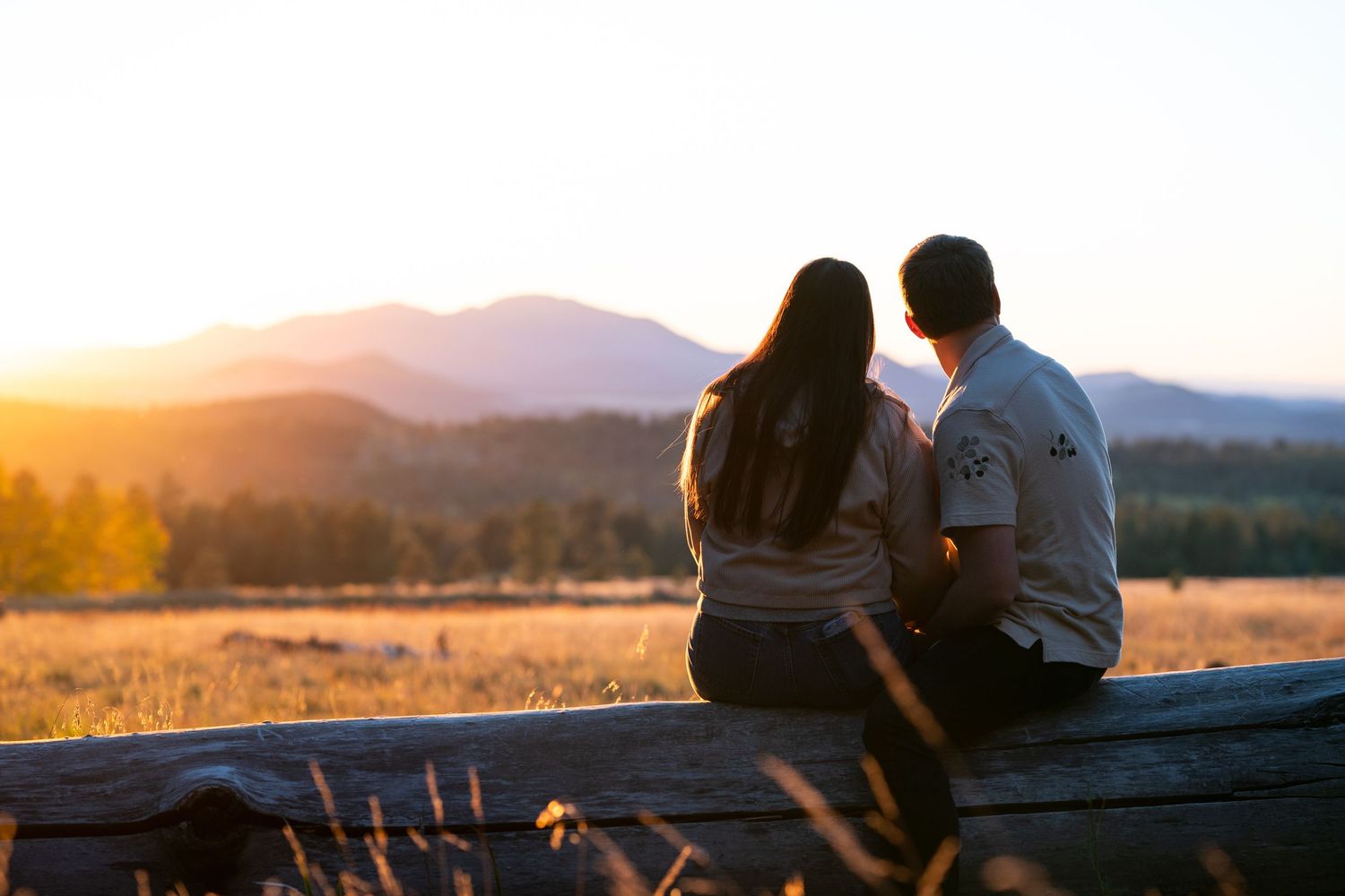 Couple sitting close on a wooden fence at sunset, gazing over a golden meadow toward silhouetted mountains and glowing sky.