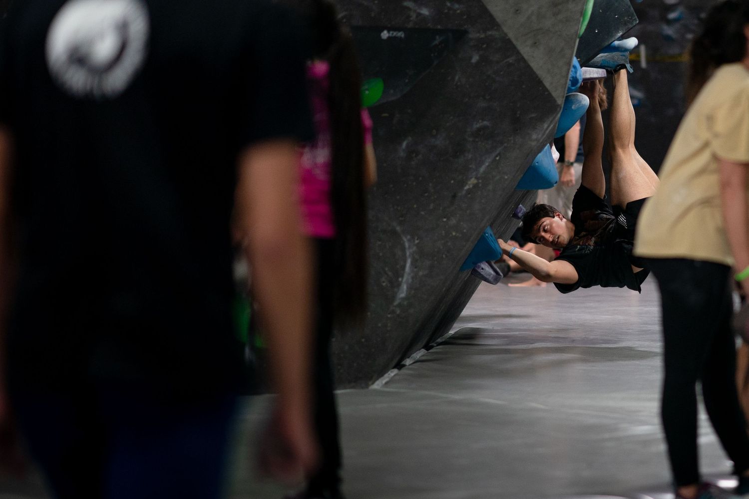 Climber hangs upside down on an overhanging indoor bouldering wall while spectators stand nearby in a climbing gym.