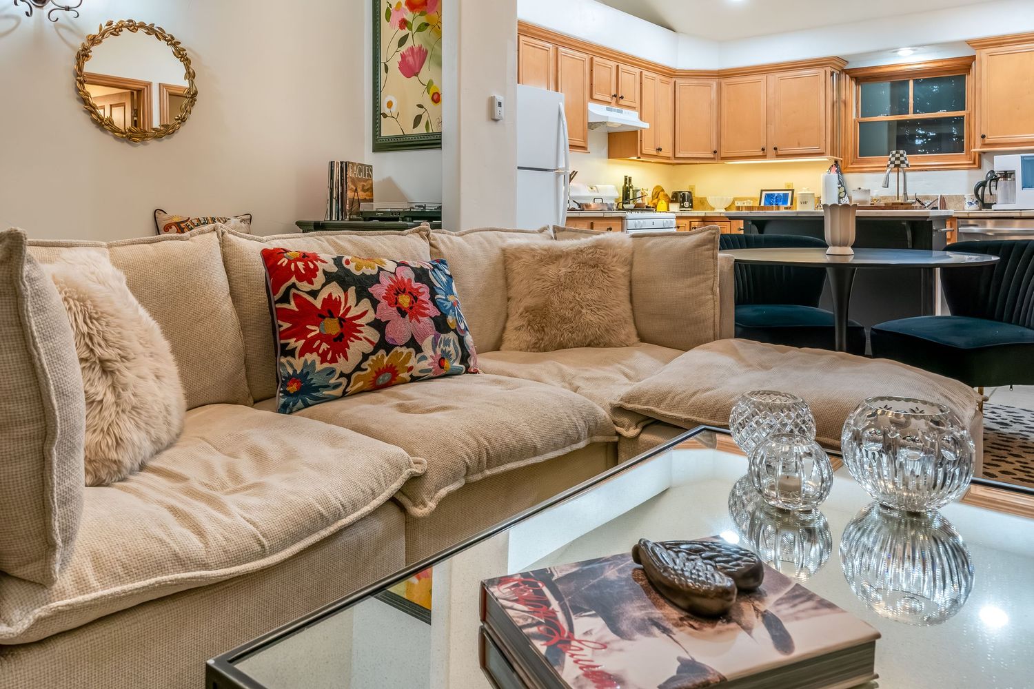 Cozy living room with beige sectional sofa, decorative pillows and glass coffee table adjacent to open kitchen area.