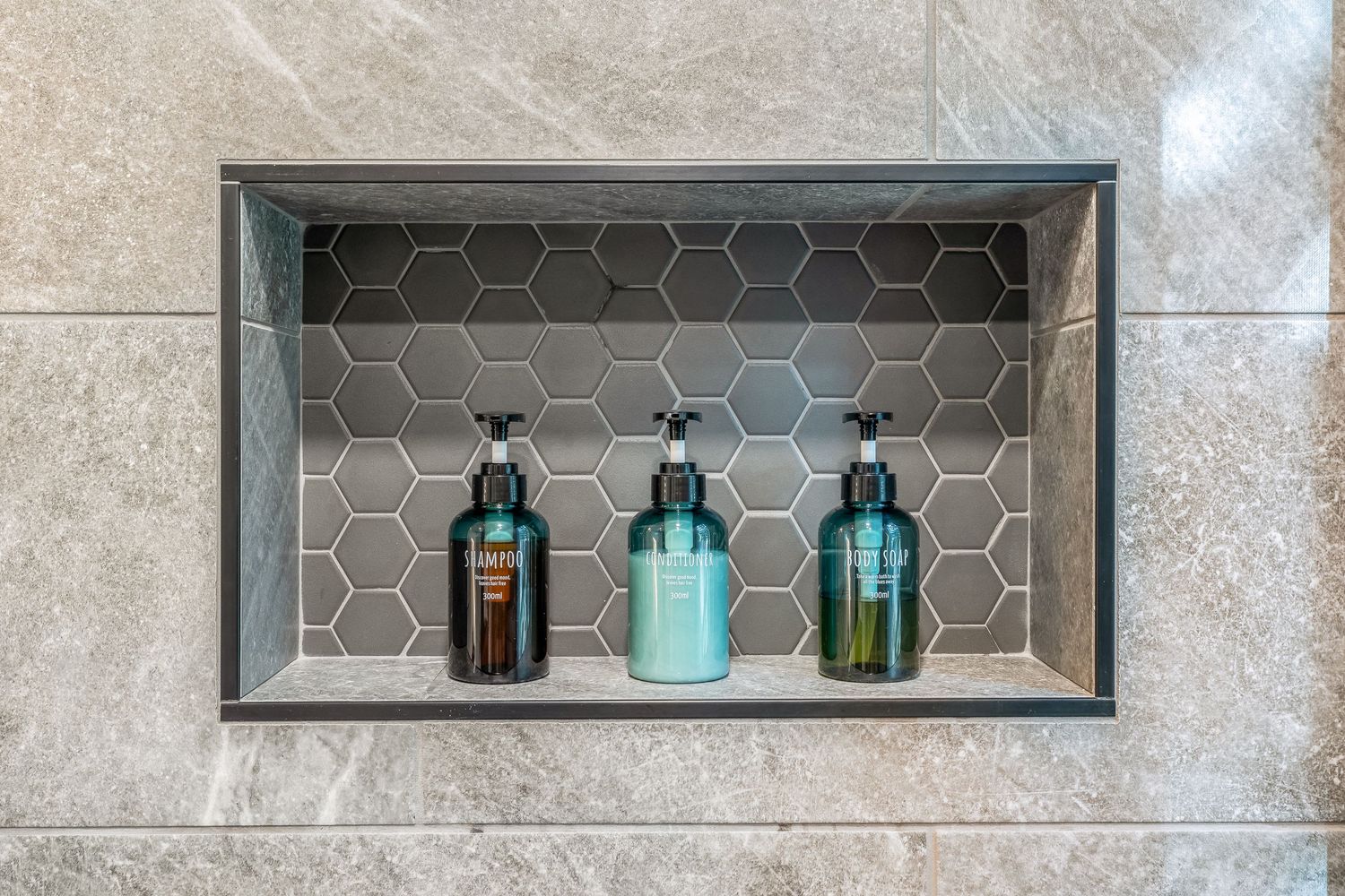 Three pump bottles on a recessed shower niche shelf with hexagonal mosaic tile backsplash in a modern gray tiled bathroom.