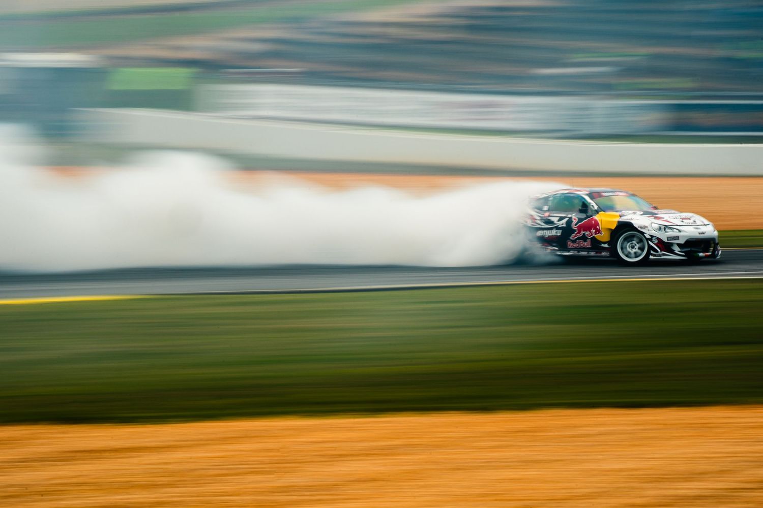A Red Bull sponsored drift car creates massive smoke clouds while sliding sideways on a racing circuit at high speed.