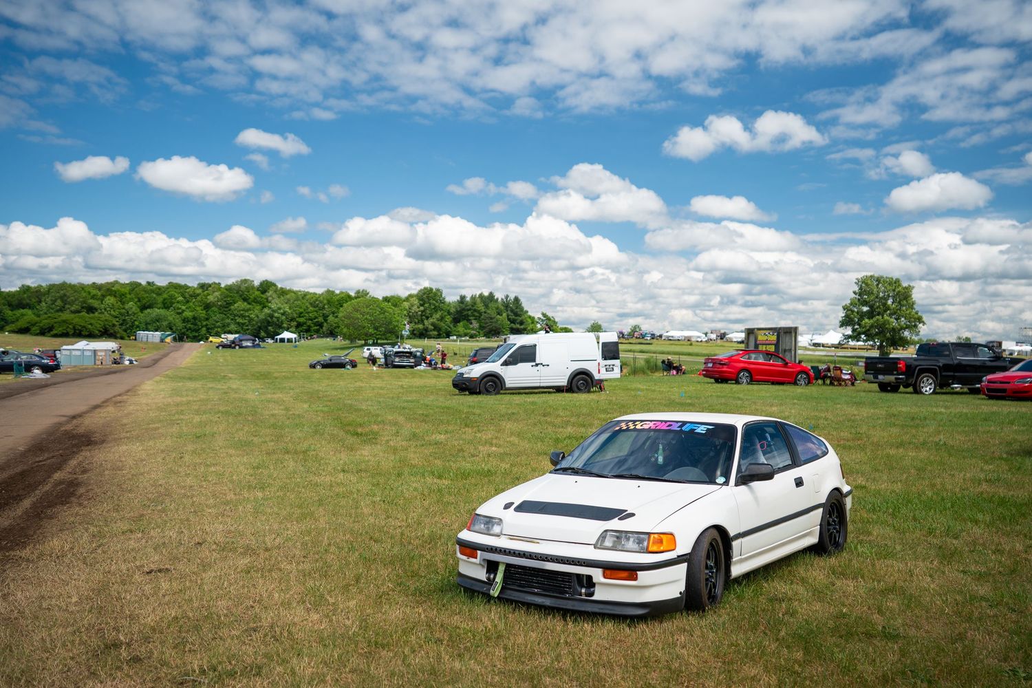 White Honda CRX race car parked on green grass at an outdoor motorsport event under blue skies.