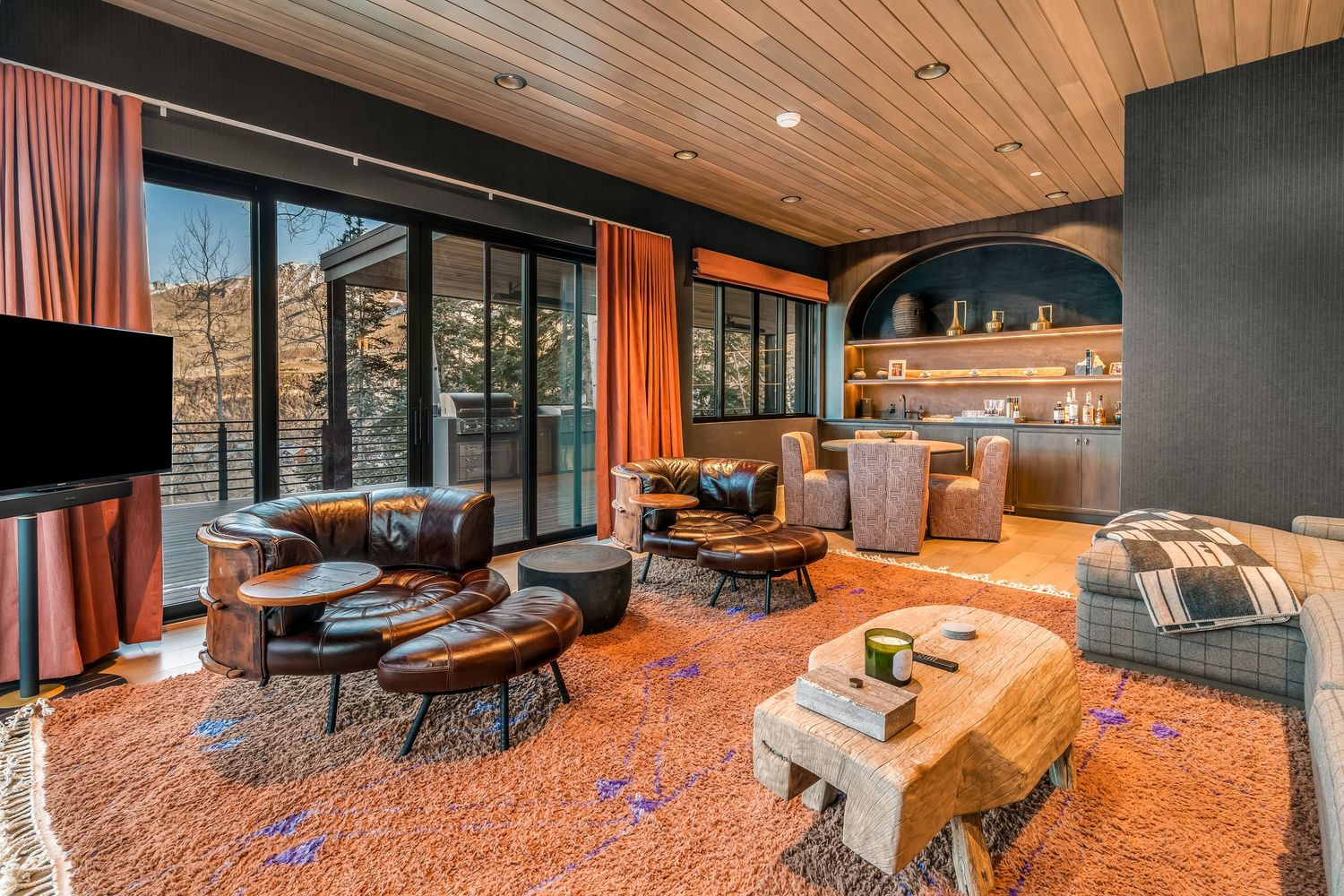 Modern living room with wooden ceiling, orange carpet, and large windows offering mountain views.