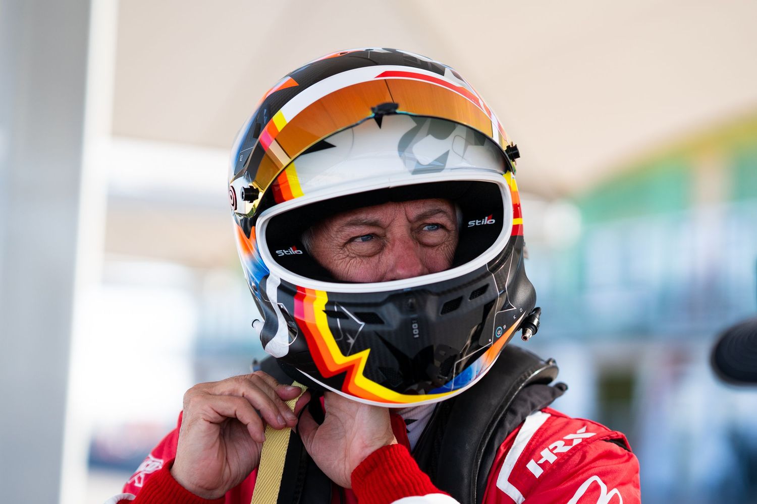 Racing driver in red uniform and white helmet making adjustments before race.
