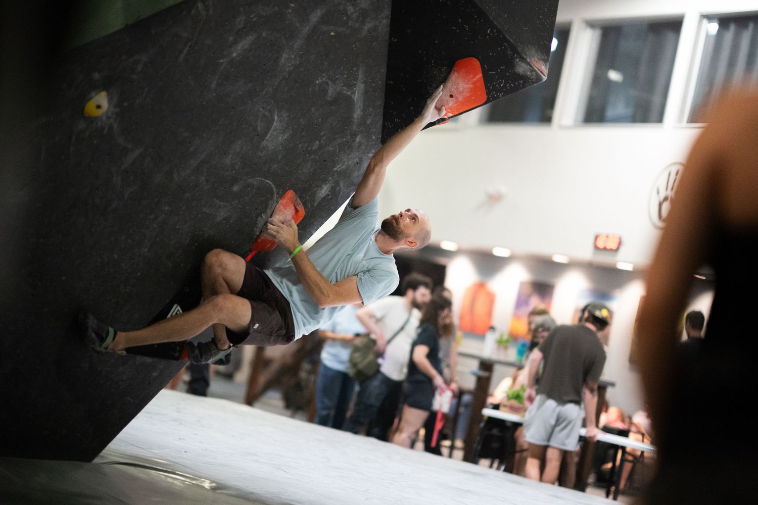 Boulderer in a gray shirt stretches for an orange hold on a dark overhanging wall in a busy indoor climbing gym.