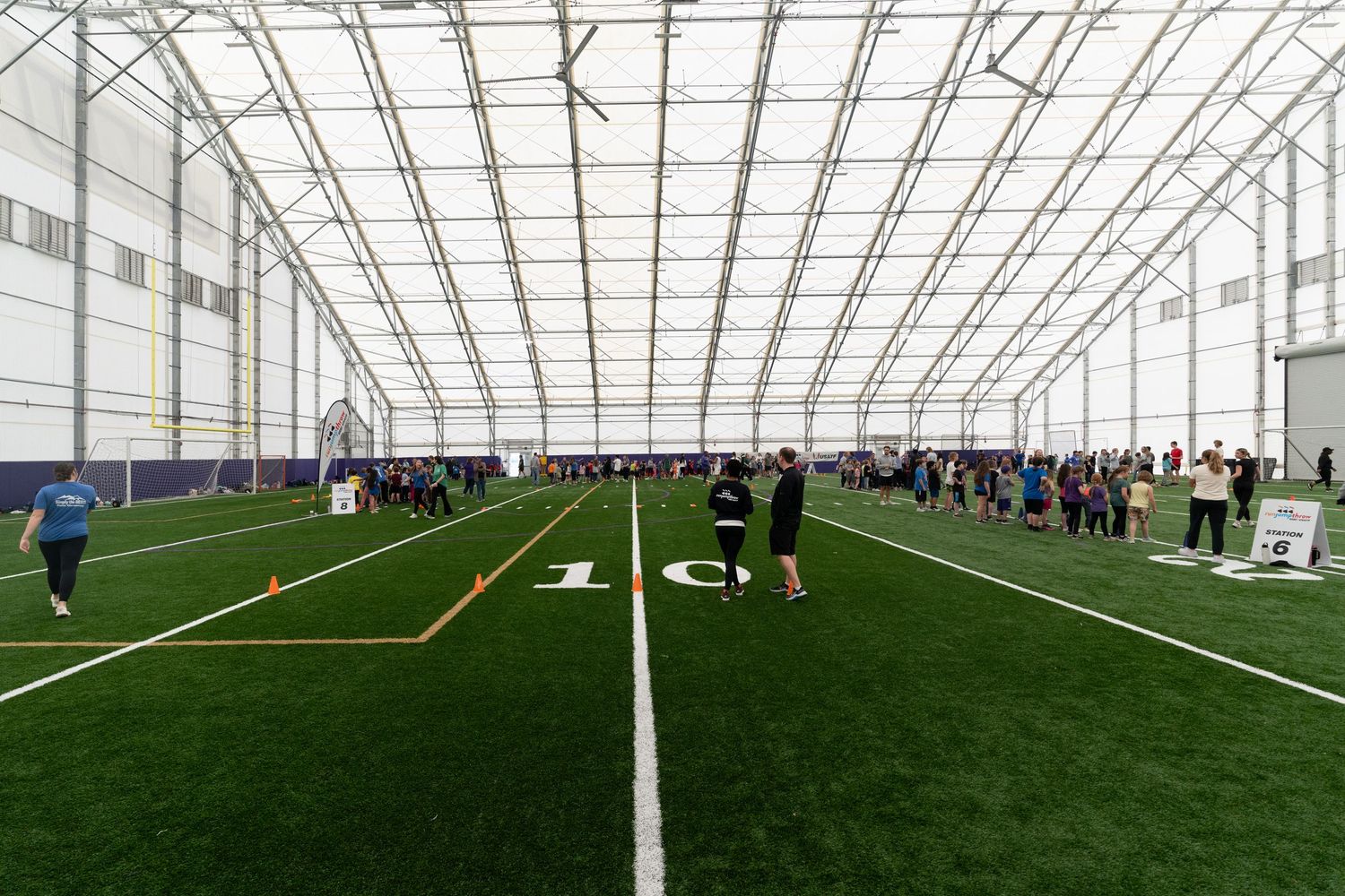 Interior view of a large indoor sports facility with artificial turf and white line markings.