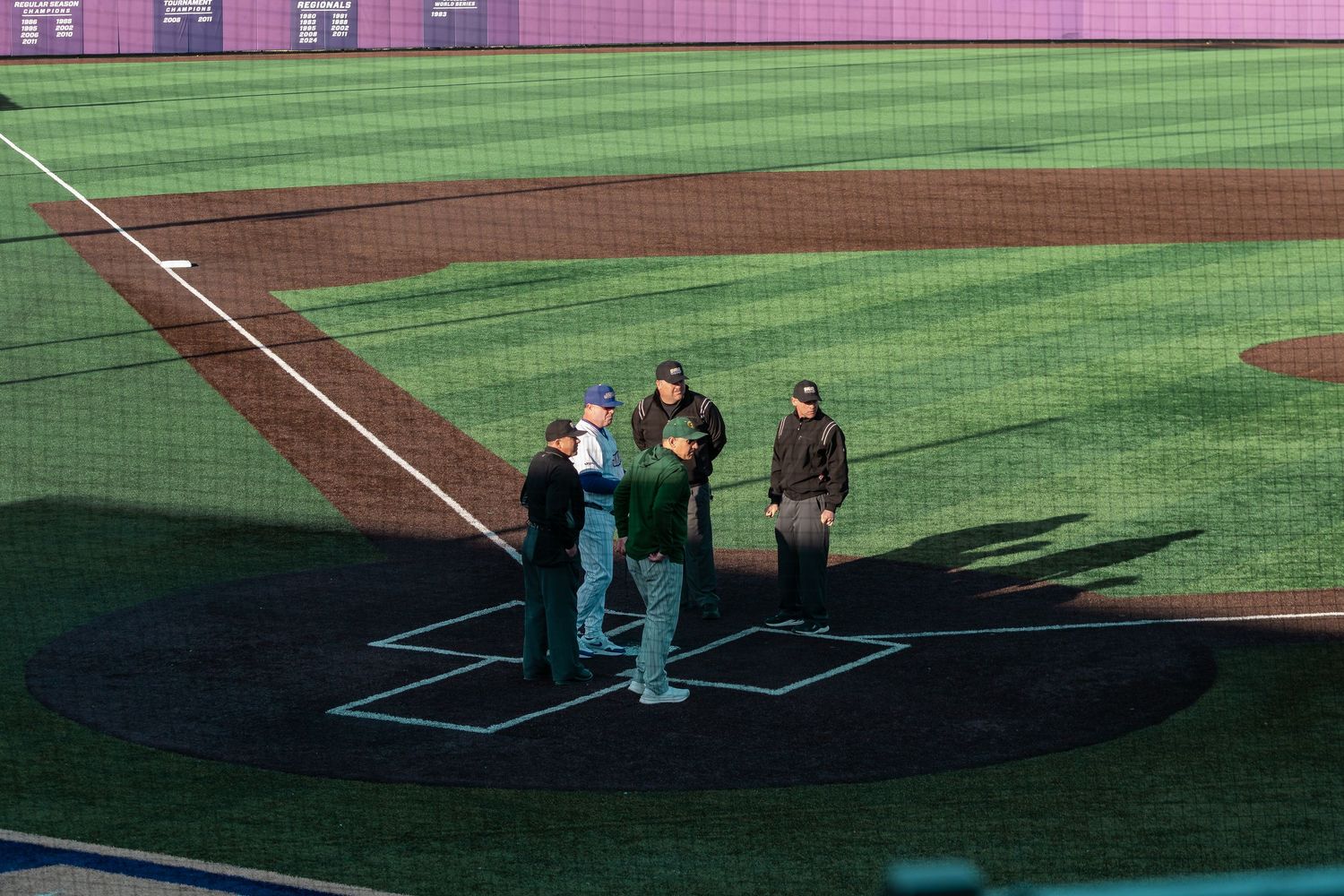 Baseball players practicing on a green field with stadium seating and pink walls visible in the background.