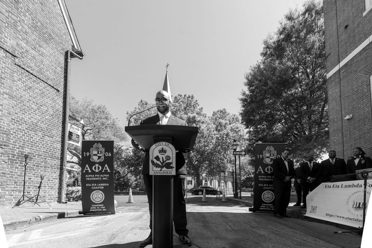 Speaker addresses audience from podium with organizational banners displayed.