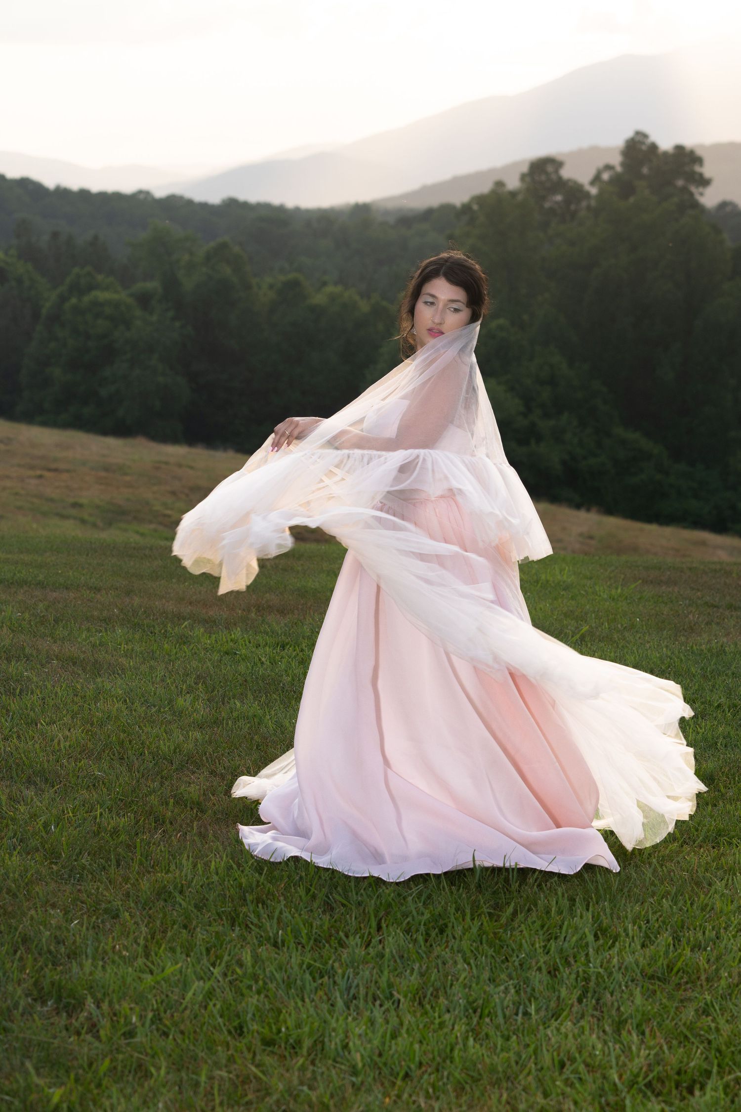 Flowing white wedding dress billows dramatically across green hillside with mountains behind.