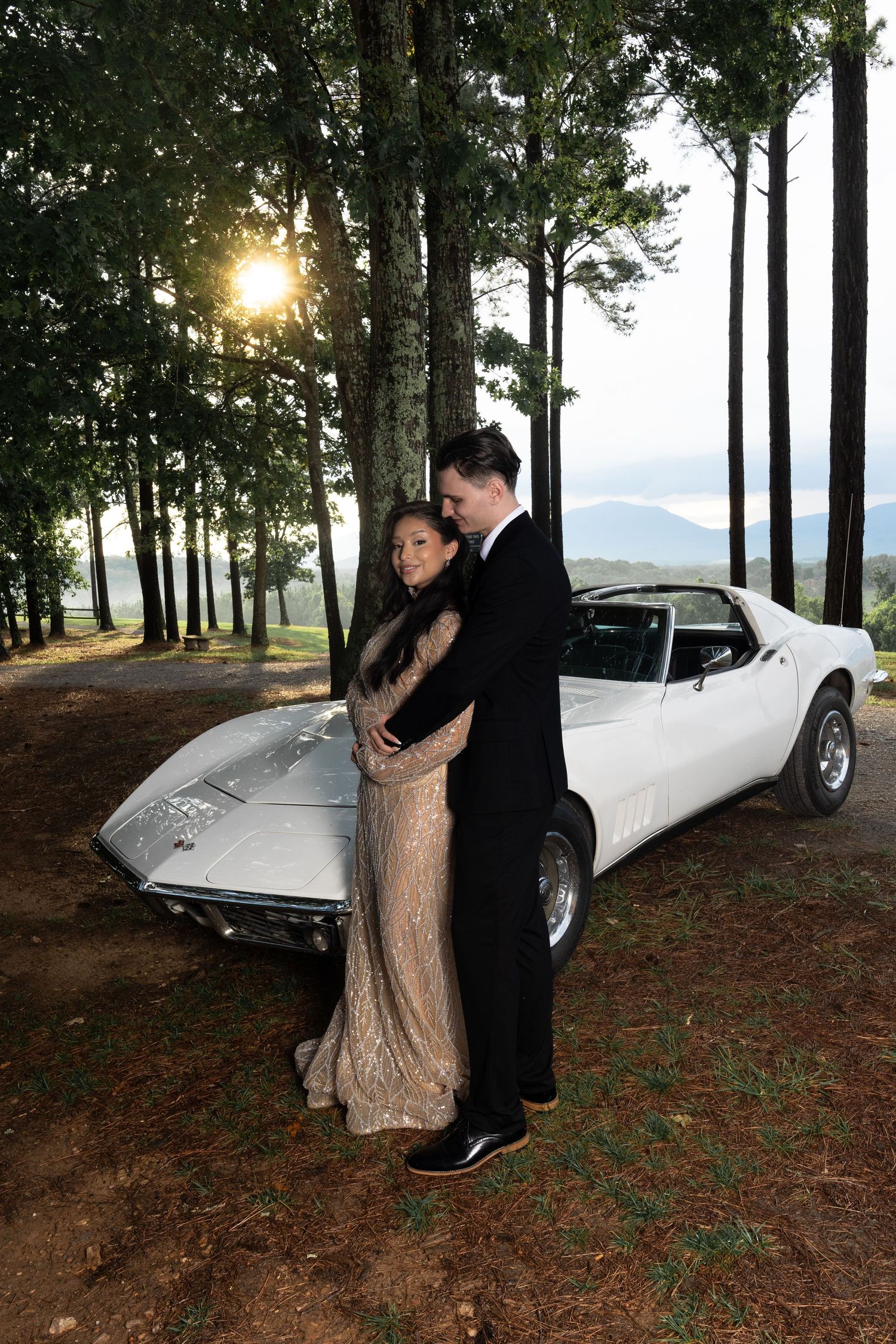Couple poses with classic white vintage sports car among tall pine trees at sunset.