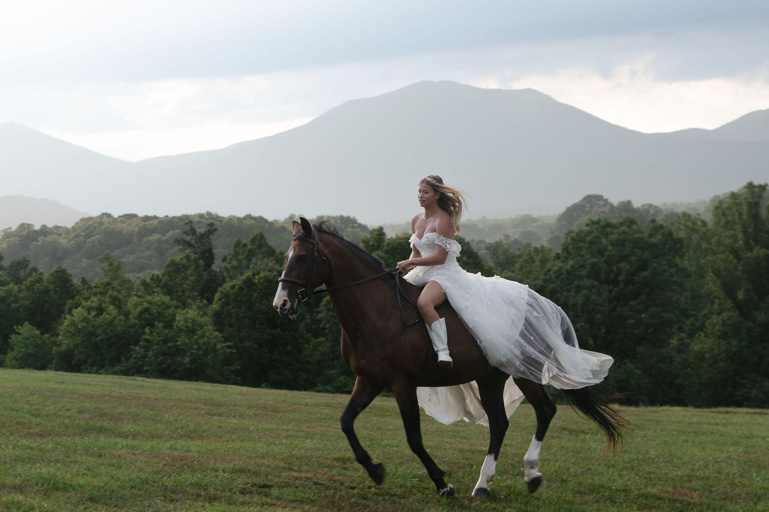 White dress flowing as person rides horseback across a scenic mountain meadow.
