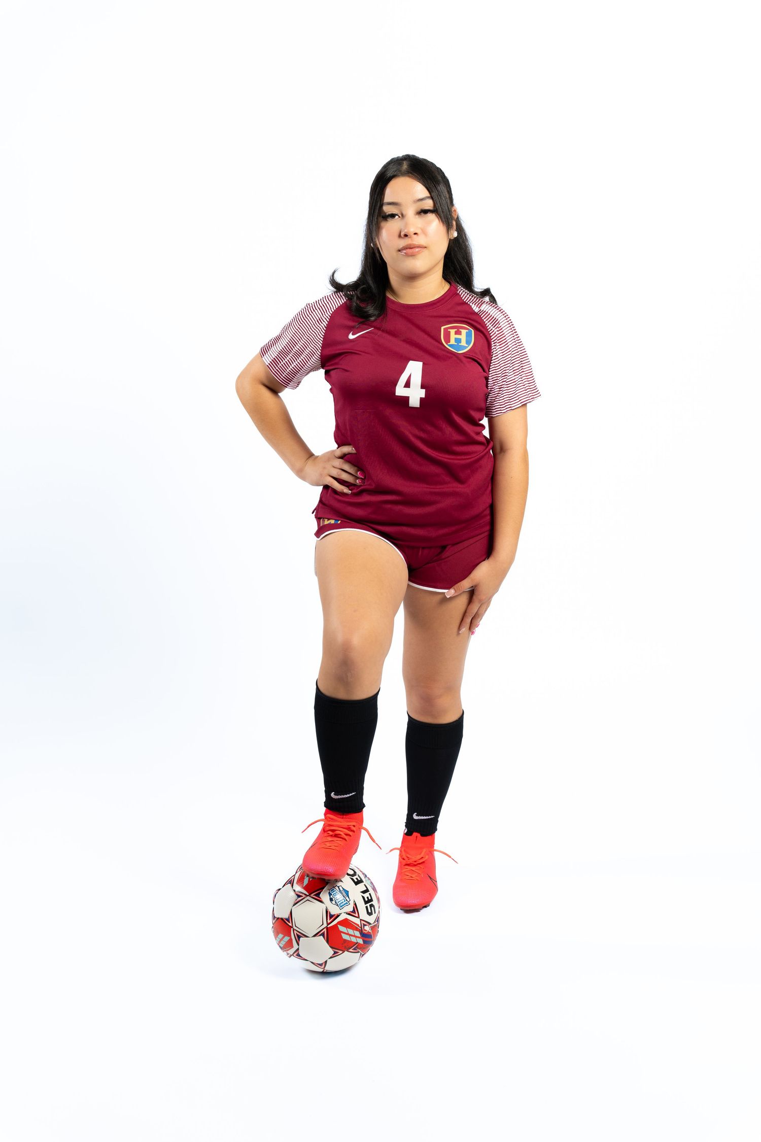 Soccer player in maroon jersey poses with ball in studio against white background.