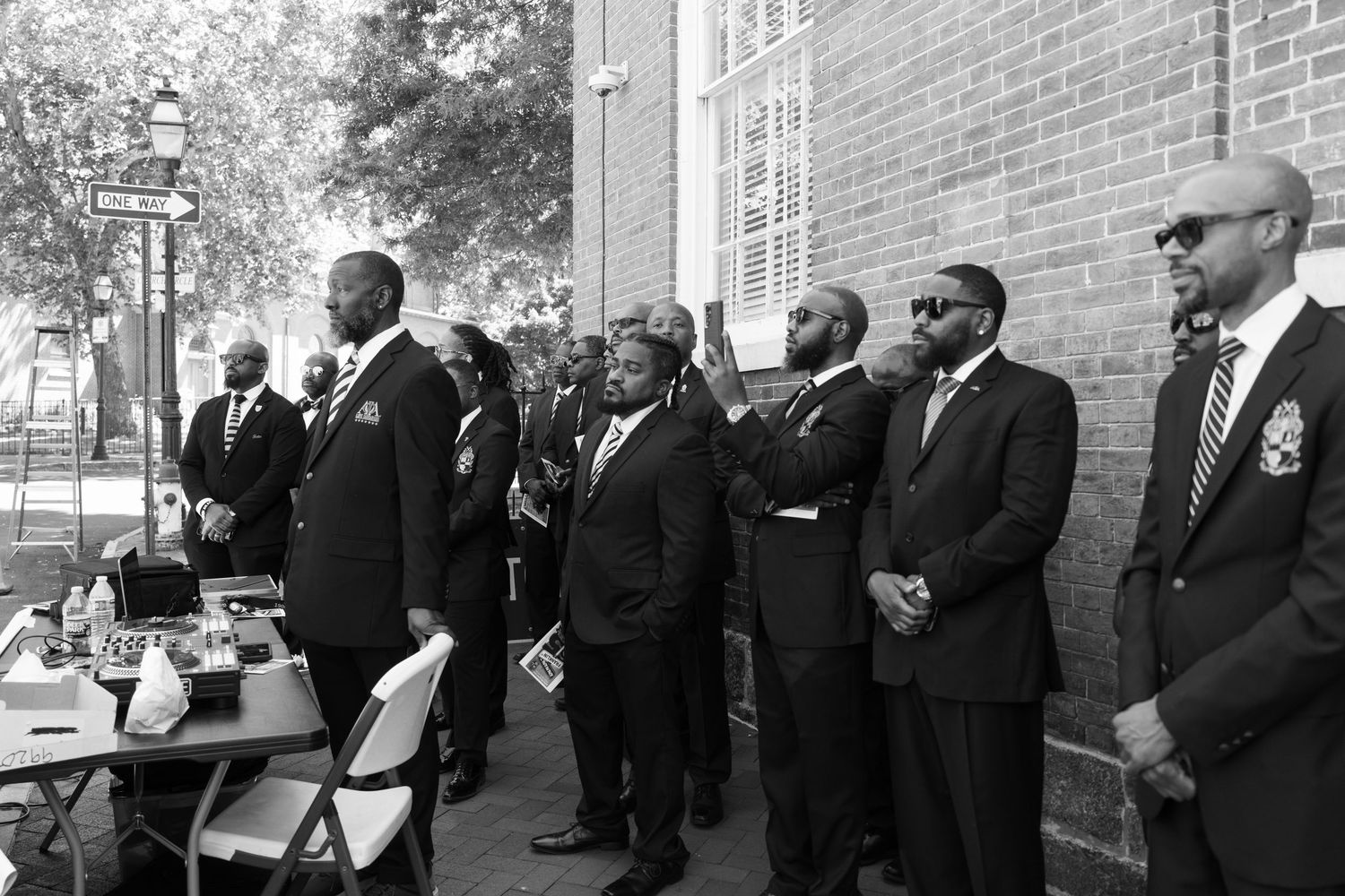 Row of formally dressed attendees stand at outdoor ceremony in black and white.