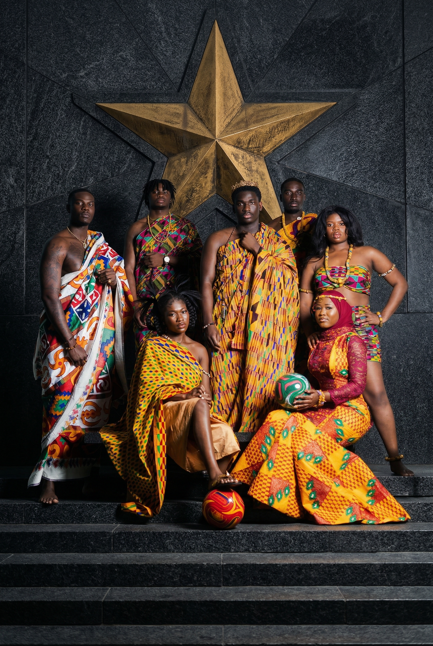 A group wearing vibrant traditional African kente cloth and ankara prints poses against a dark wall with large gold star.