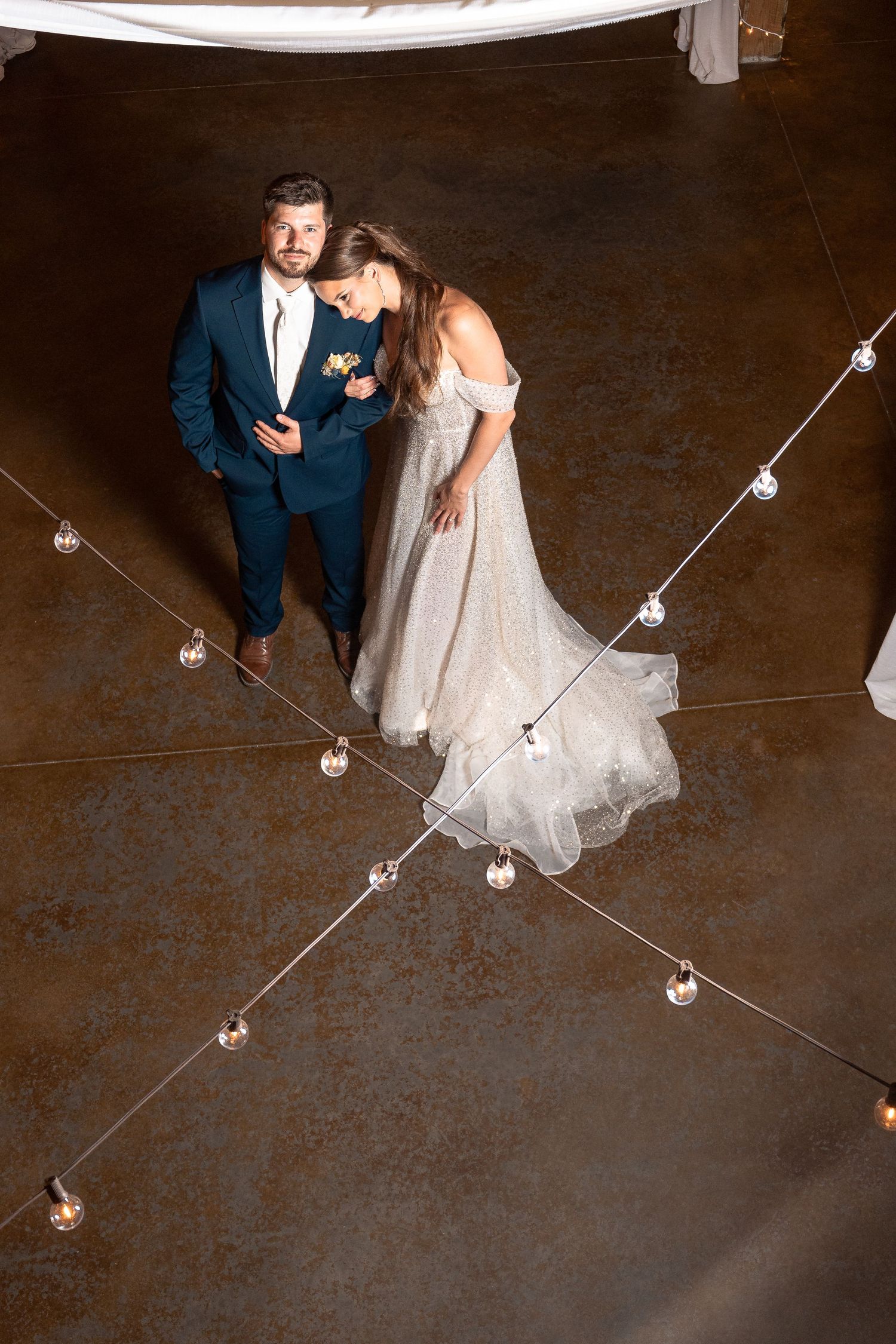 Wedding couple photographed from above on rustic floor with string lights creating geometric patterns.
