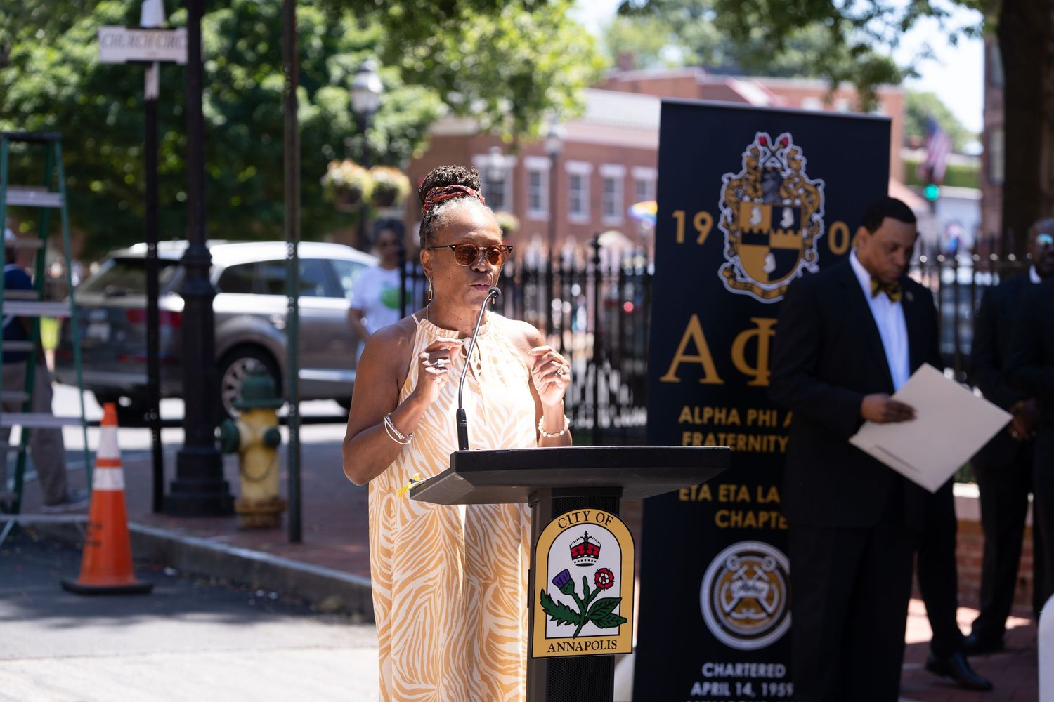 Speaker addresses audience at outdoor podium with Alpha Phi Alpha organization banner.