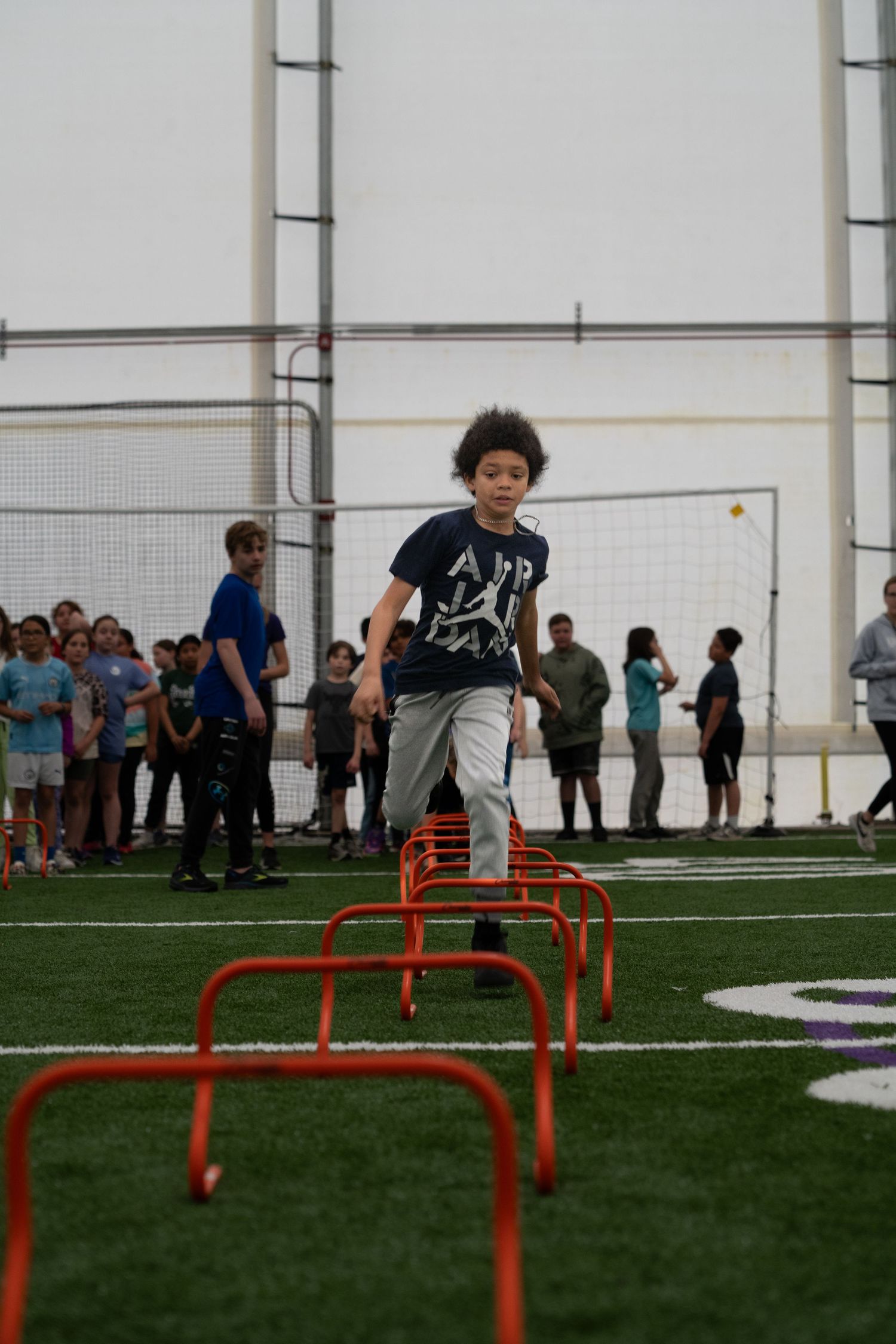 Athletes practicing hurdle jumps on an indoor athletic field with spectators watching.