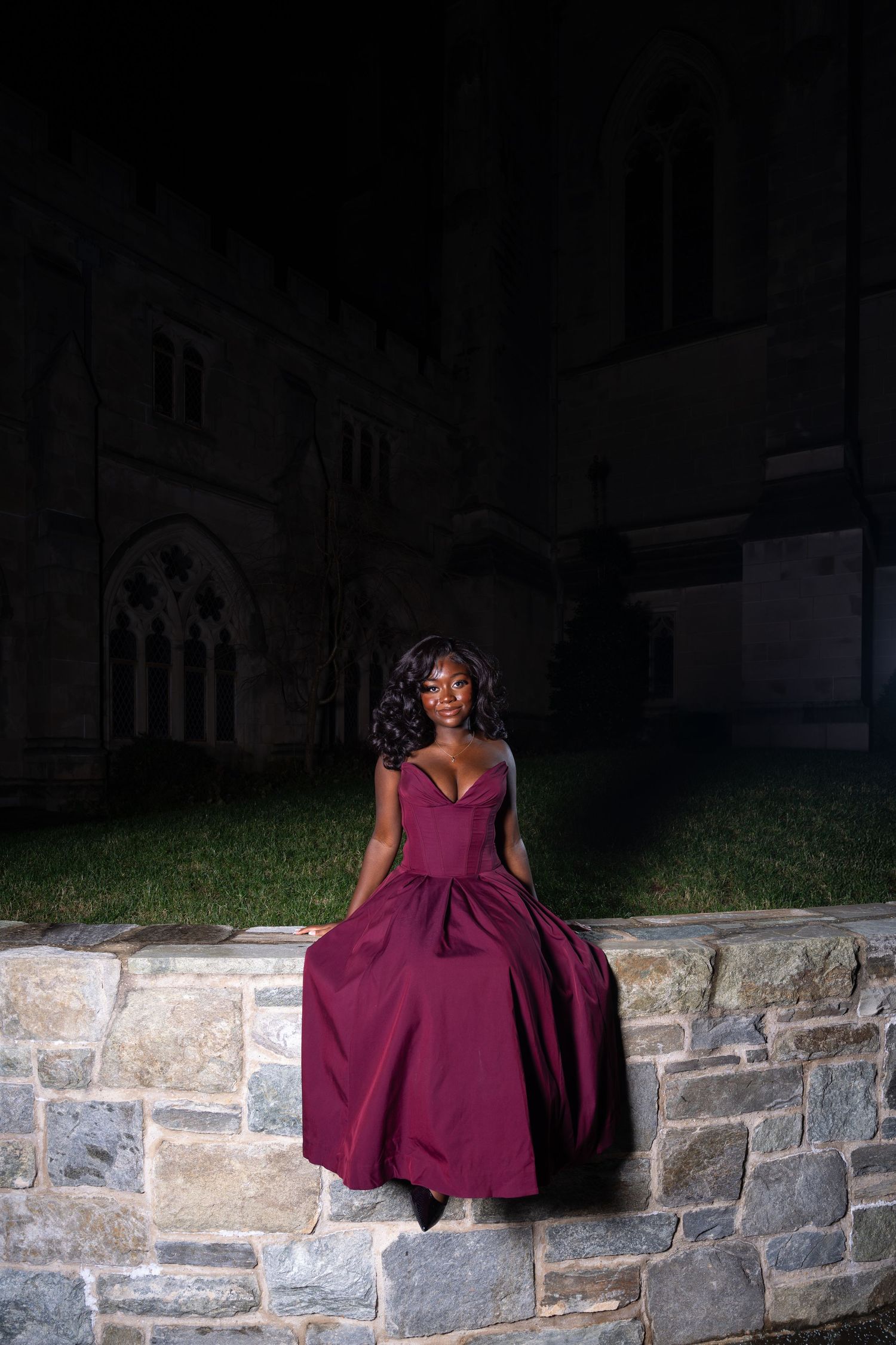 Person in burgundy dress sitting on stone wall at night with gothic architecture backdrop.