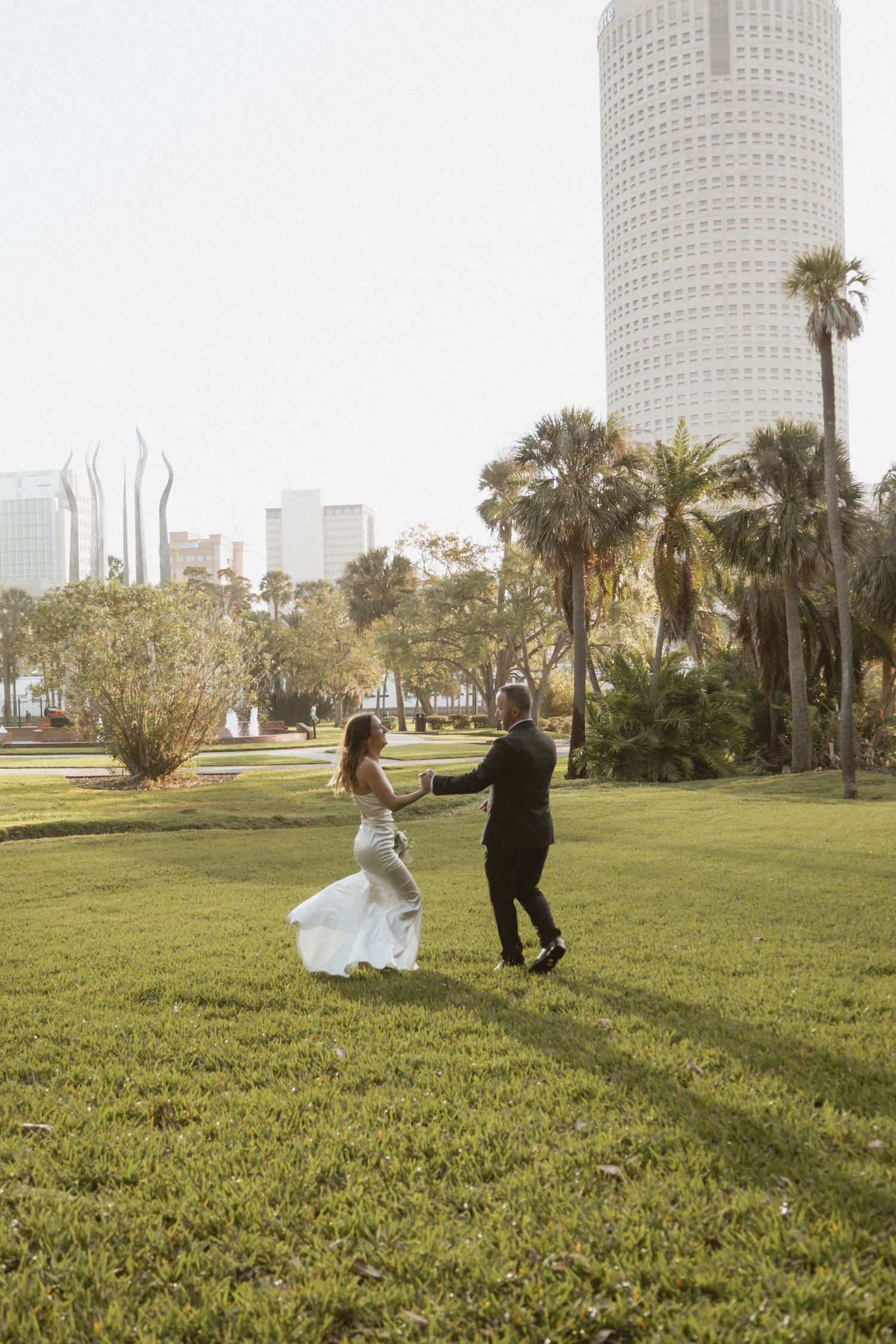 A couple in wedding attire run and play together on a grassy lawn with tall buildings in the background.