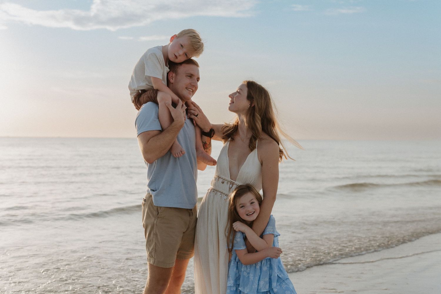Happy family of four enjoying a sunny beach day, with dad carrying son on shoulders and mom hugging daughter by the ocean.
