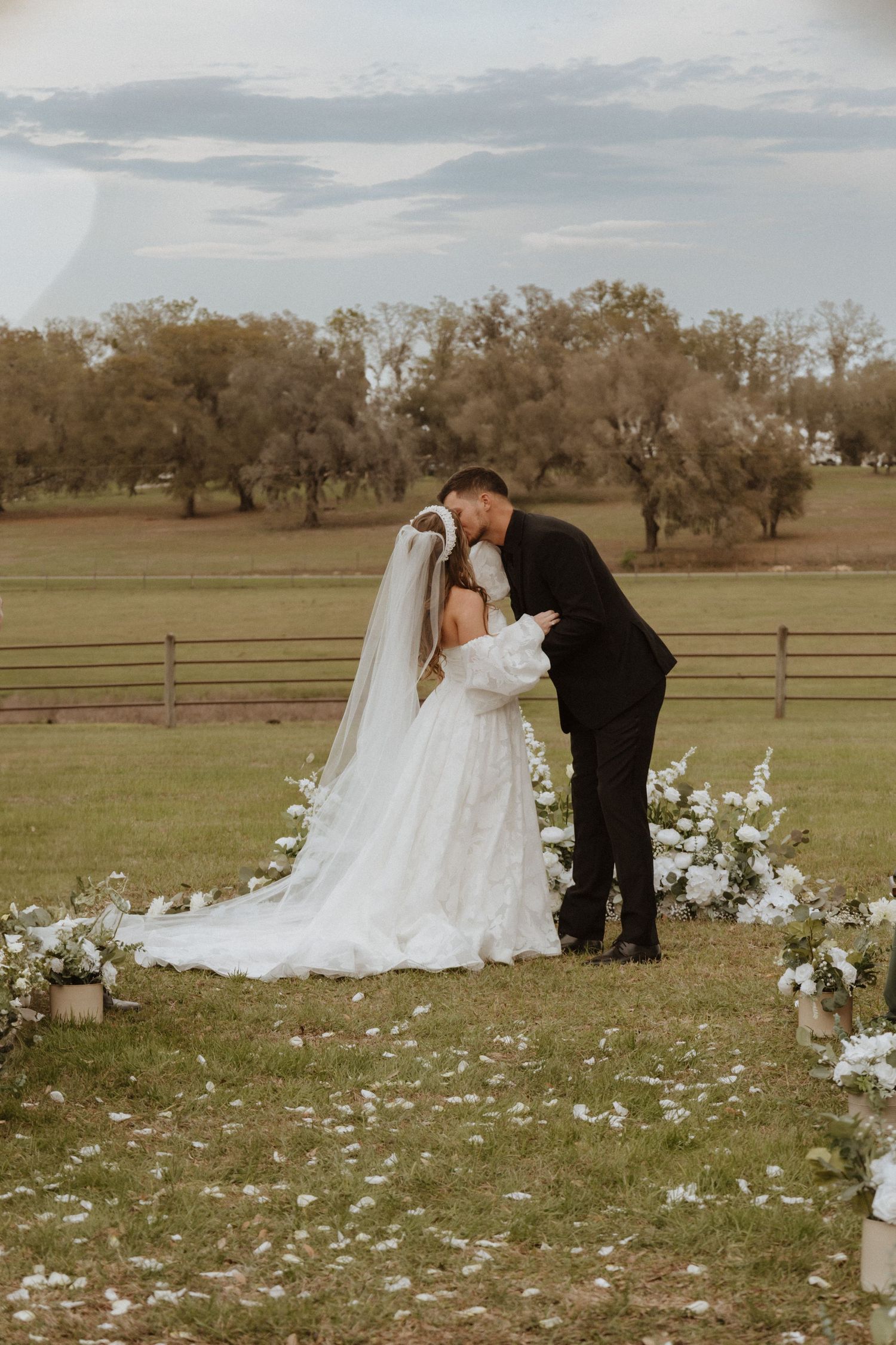 A couple shares their first kiss as newlyweds in a rustic outdoor setting with floral decor.