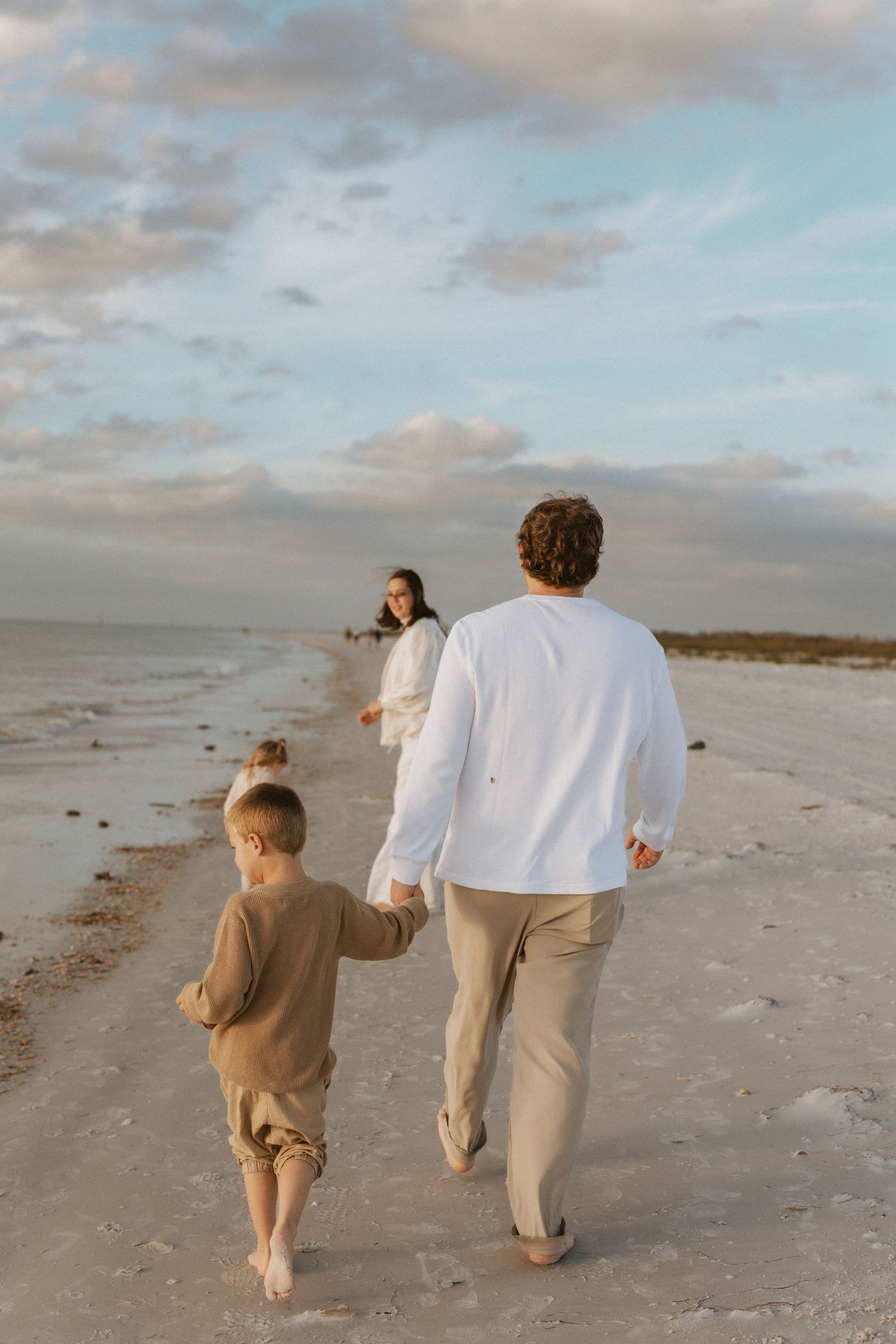 A family walks barefoot along a peaceful beach at sunset with cloudy skies overhead.