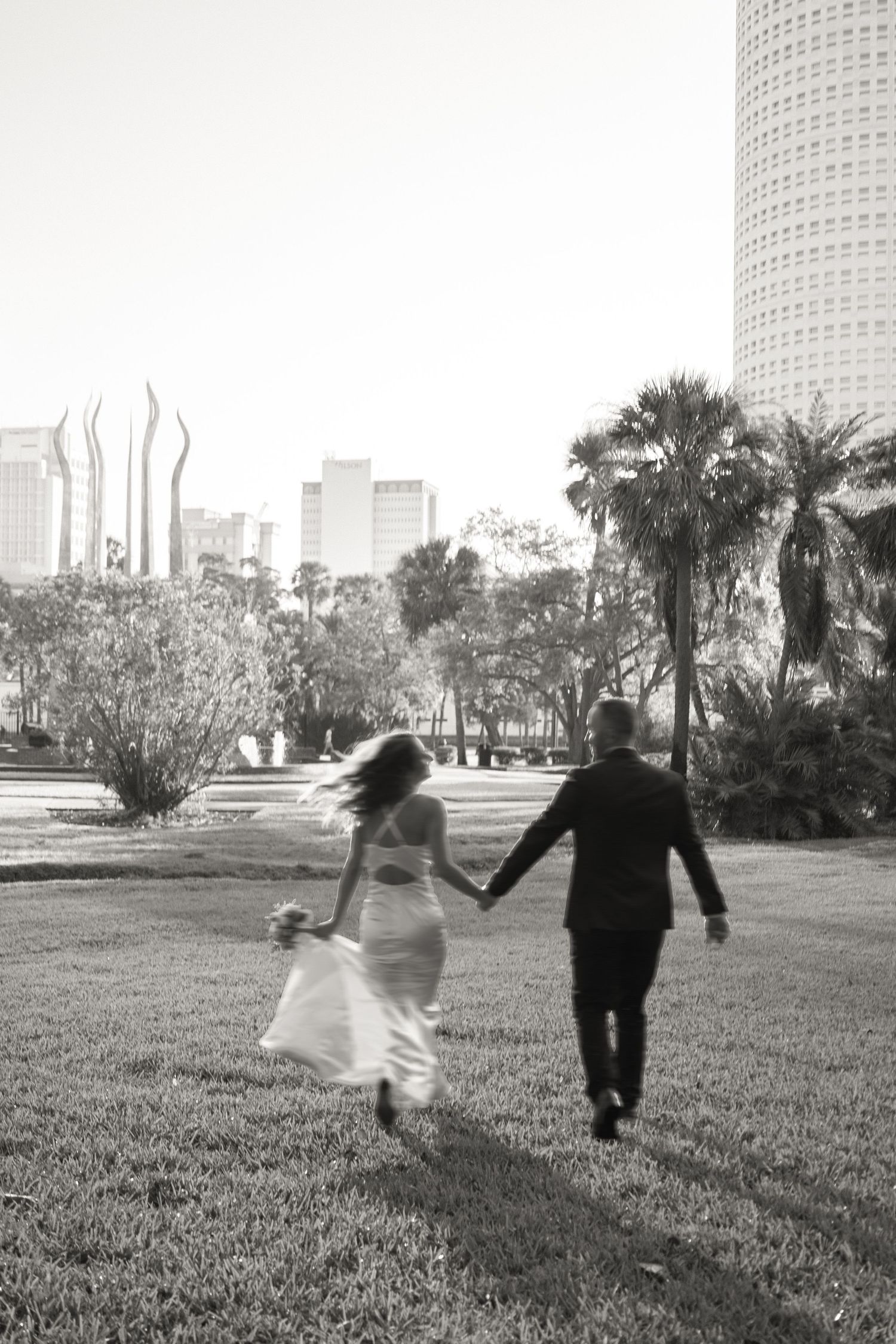 Black and white photo of a couple running playfully across a grassy field with city buildings in background.