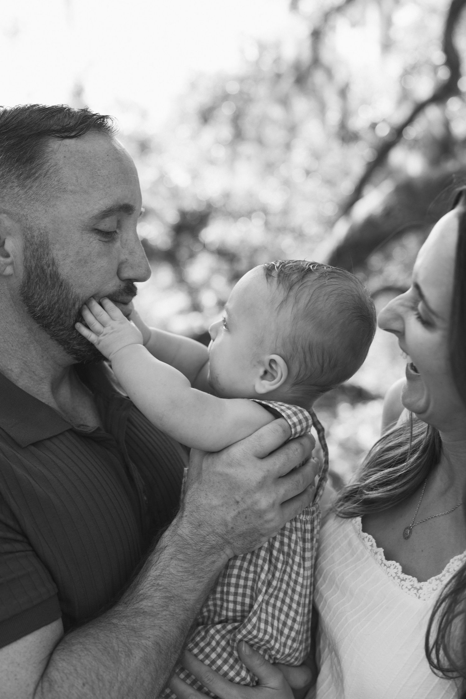 Black and white photo of parents lovingly gazing at their baby outdoors, capturing a tender family moment.