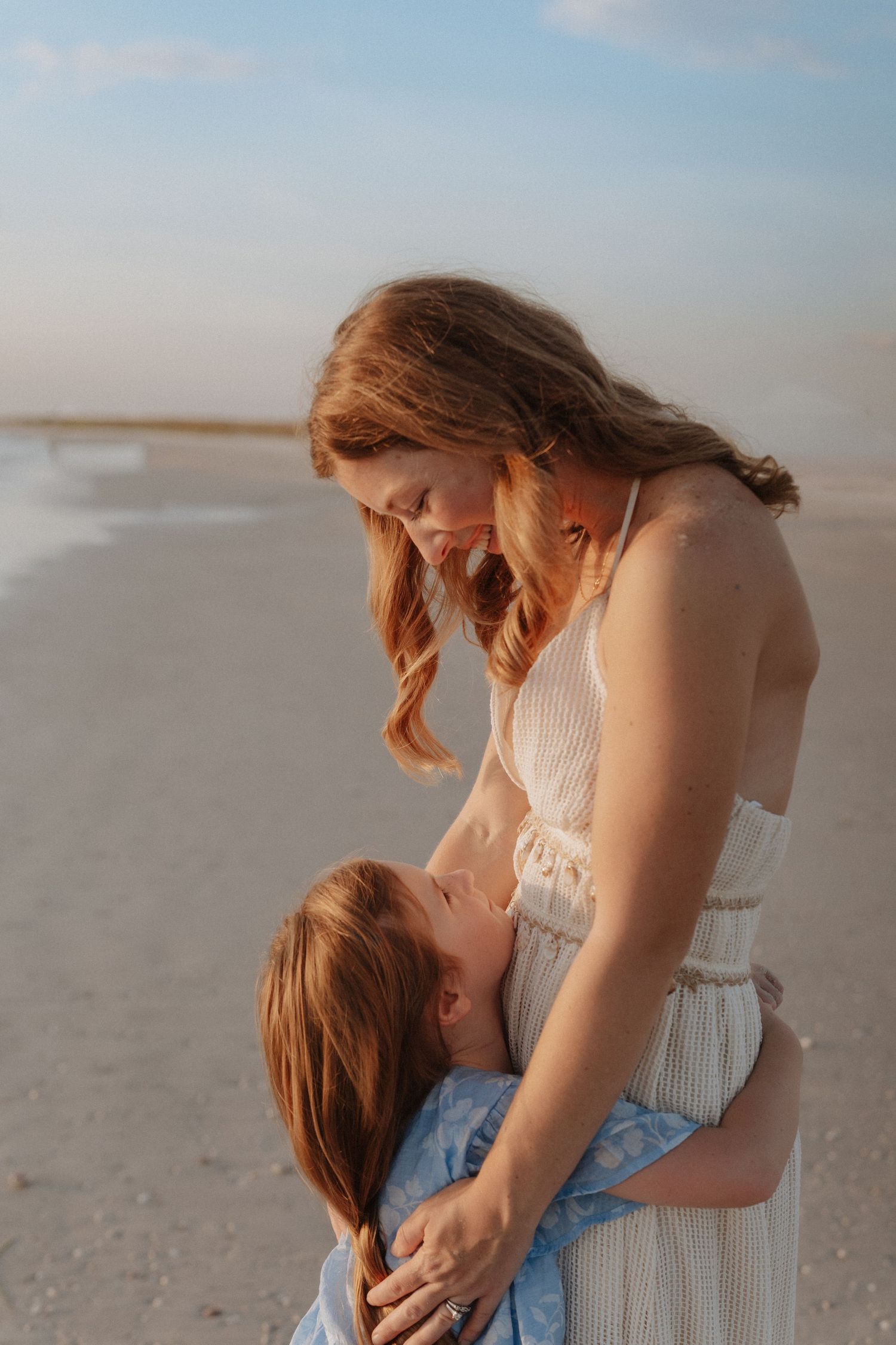 A mother and daughter share a tender embrace on a beach at golden hour, both with auburn hair.