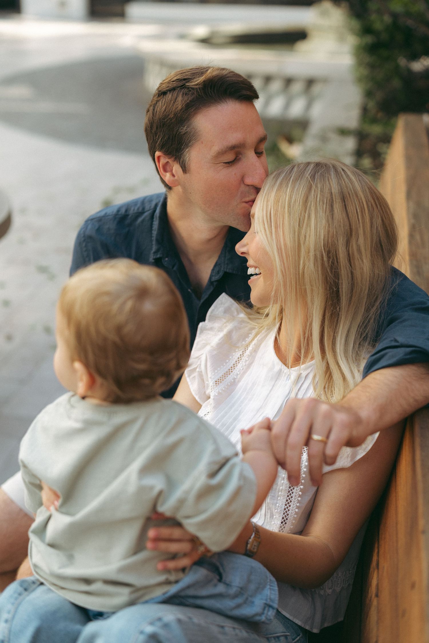 A happy couple shares a kiss while holding their baby outdoors on a sunny day.