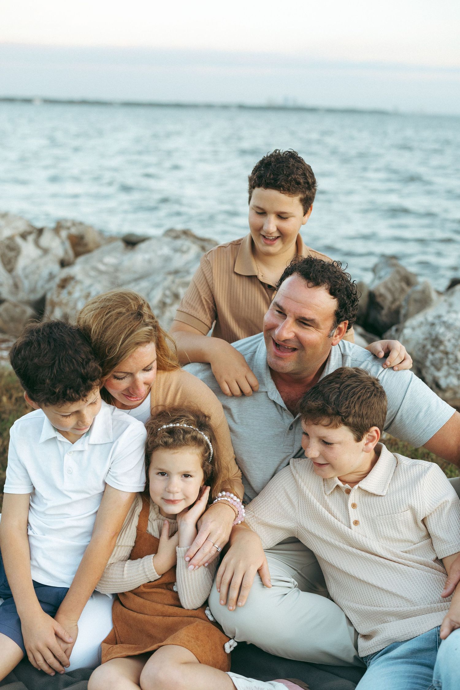 A happy family wearing white and beige clothing sits together on coastal rocks by the ocean on a sunny day.
