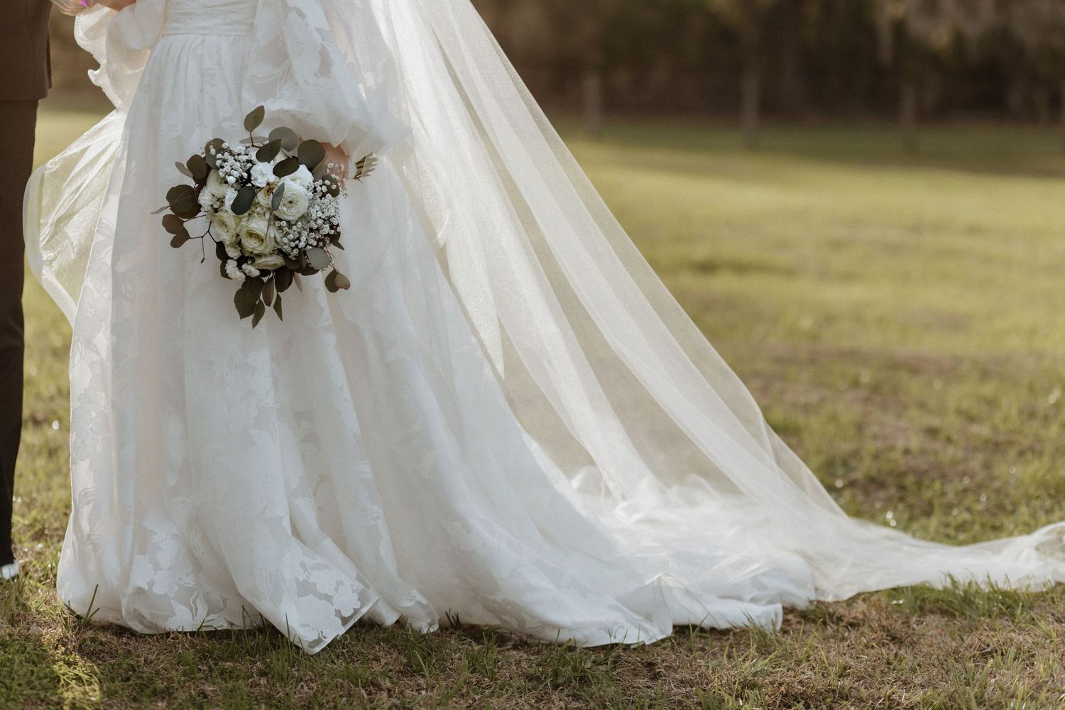 The train of a white wedding dress trails beautifully across green grass.