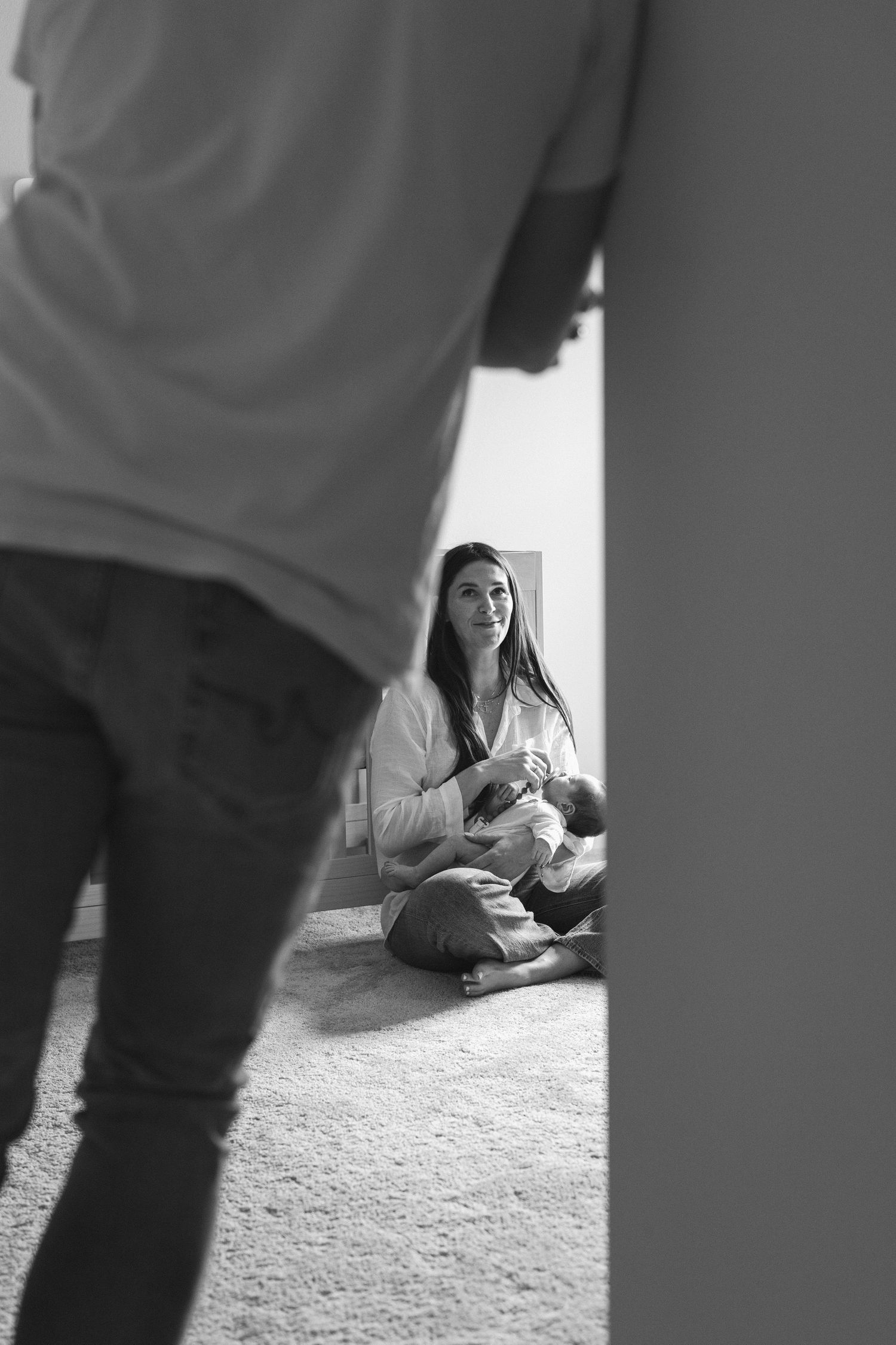 Black and white photo of a woman sitting cross-legged on floor nursing a baby, viewed through a doorway.