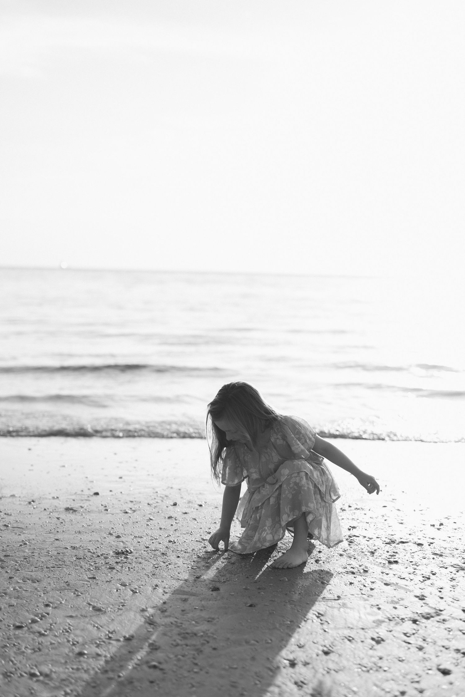 Black and white photo of a young girl in a dress crouching on a sandy beach near ocean waves.