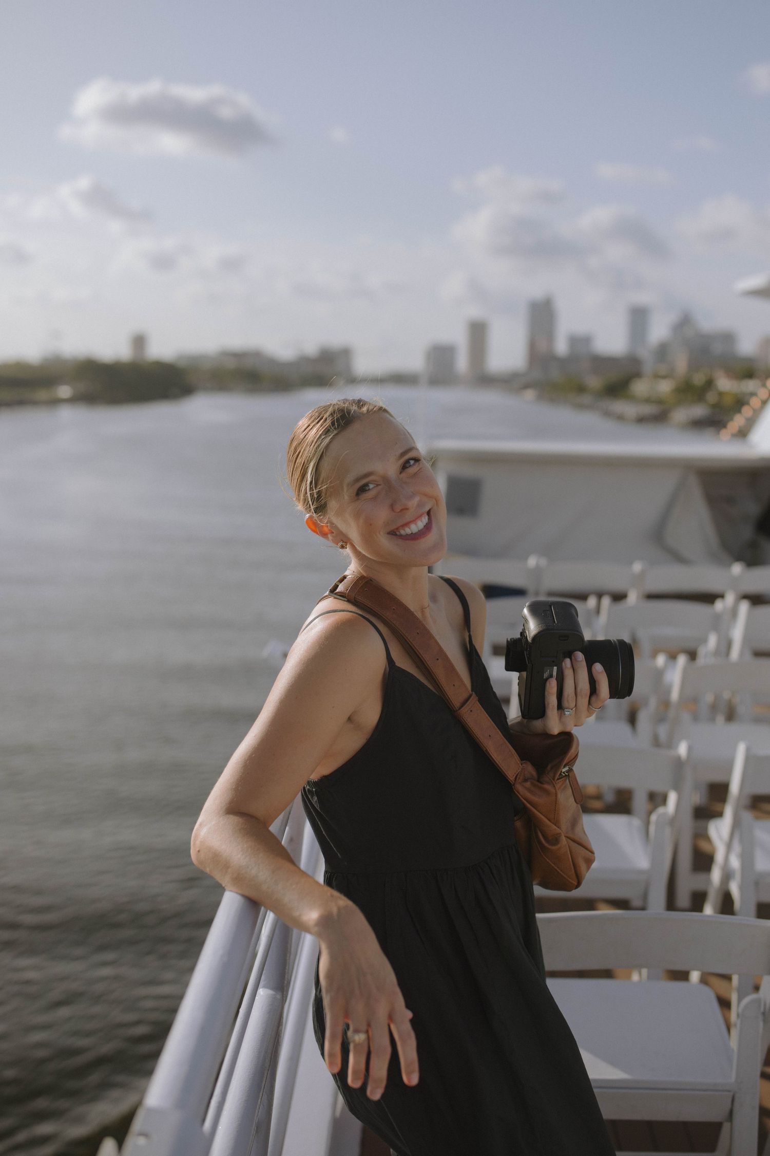 Photographer with camera stands on boat deck overlooking waterfront cityscape with tall buildings in background.