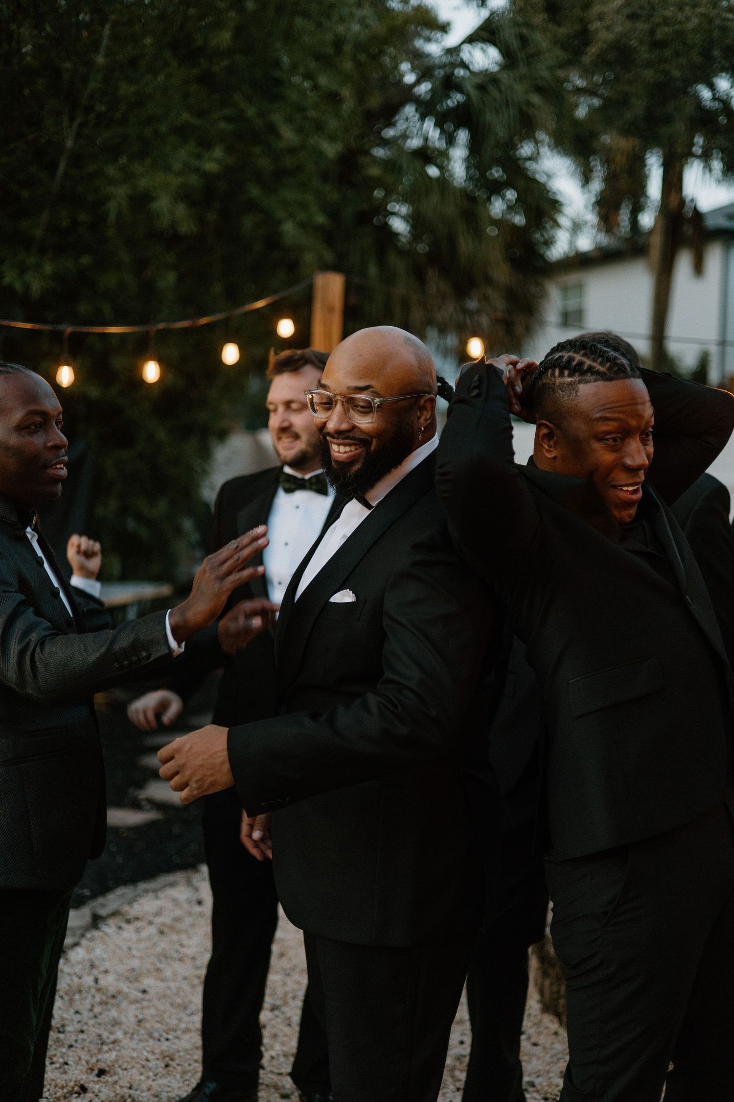 Groomsmen in black tuxedos sharing a candid moment at an outdoor evening wedding reception with string lights.