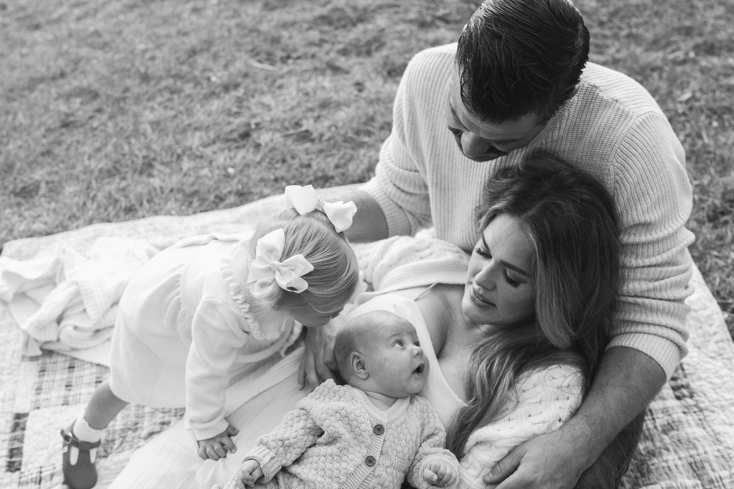 A family relaxes together on a blanket outdoors in black and white, sharing an intimate moment.