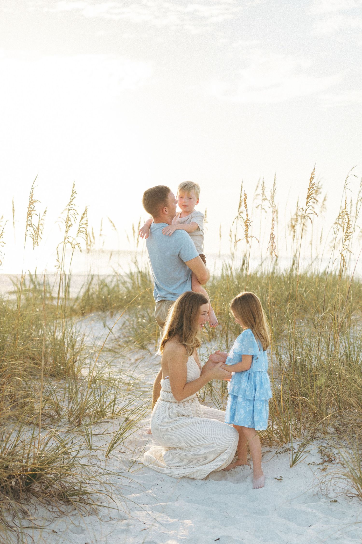 Family of four enjoying a golden hour beach photoshoot among sea oats on sandy dunes, dressed in coordinating blue and white outfits.