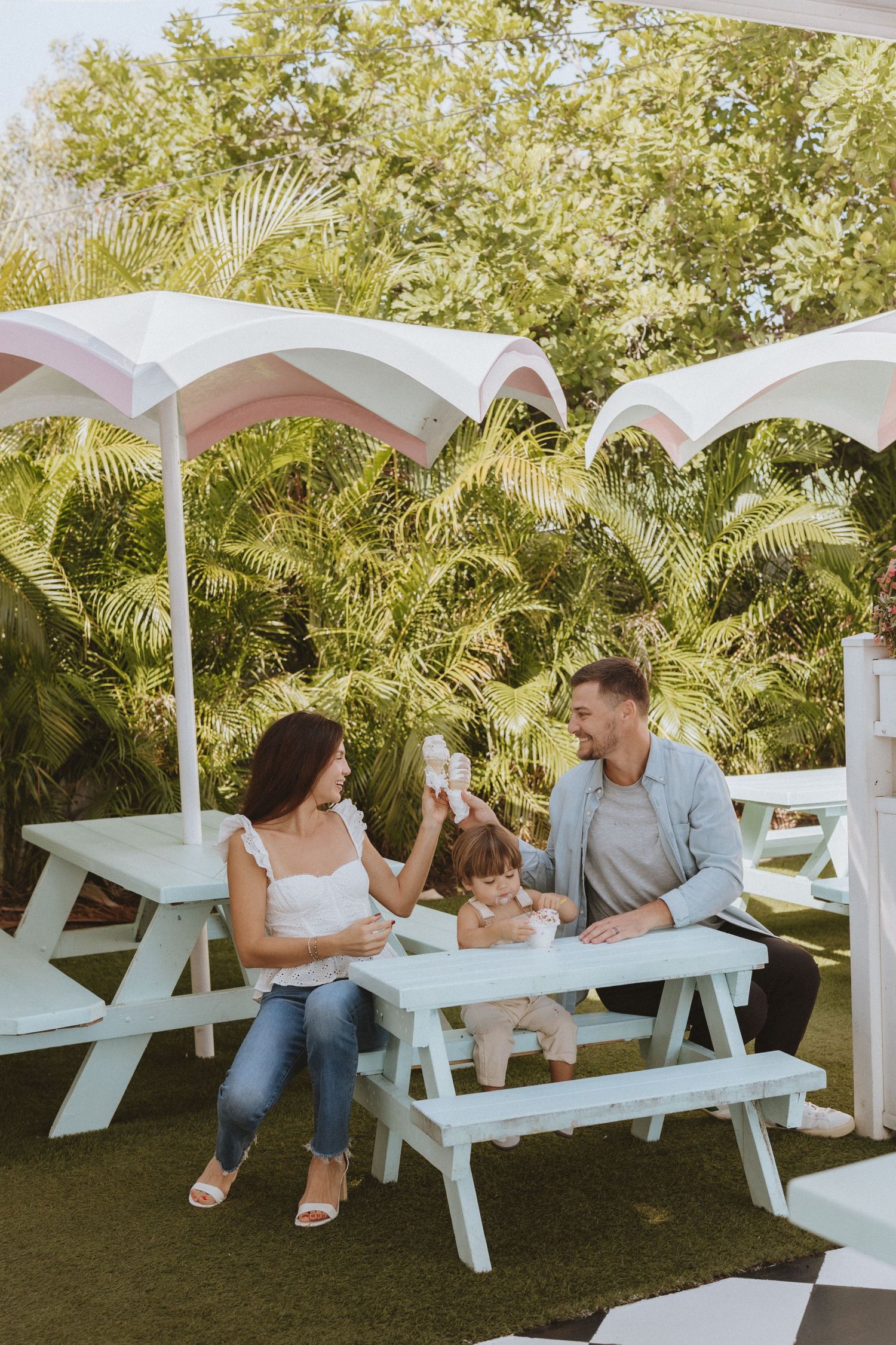 A family sits together on white picnic benches under white umbrellas in a lush garden setting.